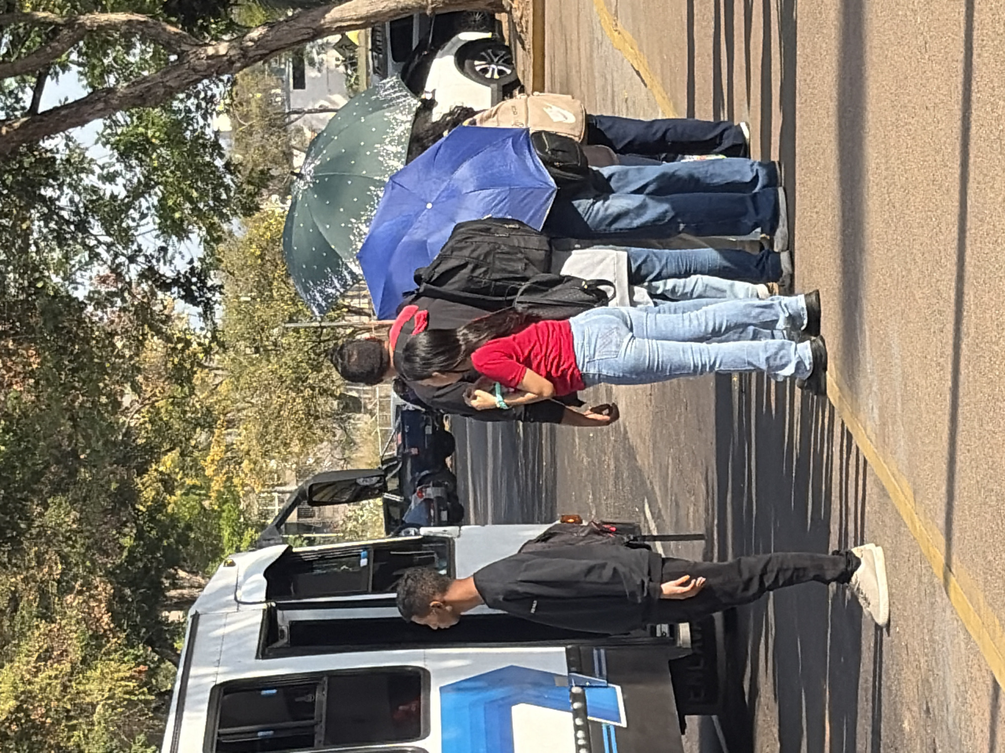 This image, set in Ciudad Guayana, Venezuela, captures an outdoor scene on a bright, sunny day, featuring several people and a bus. The photo is rotated 90 degrees clockwise.
In the foreground, a man in a black jacket, black pants, and white shoes stands beside a white bus or minibus, facing away from the camera. The bus has a blue design or logo visible on its side, with the partial text "CALV" also visible.
Behind the bus, a group of people is queued in a line. A woman in a red shirt and blue jeans is prominent in the line, looking down at a phone she's holding. Behind her, a man in a black shirt is also looking at a phone. Several other individuals are visible further down the line, mostly their legs in various shades of blue jeans. Many in the queue are carrying bags or backpacks, including a black backpack on the man in black and a beige bag over a shoulder further back. Two umbrellas are open in the queue: one dark green with white dots, and one solid blue, suggesting a need for shade from the strong sun.
The background features lush green foliage and trees, indicating a natural or park-like setting adjacent to the road. A white car is partially visible behind the people. The ground is asphalt or concrete, with a yellow line marking what appears to be a curb or edge of a pathway on the right. Long, distinct shadows on the ground and against the wall on the right indicate strong midday or early afternoon sunlight. The overall activity appears to be people patiently waiting to board the bus or for transport.