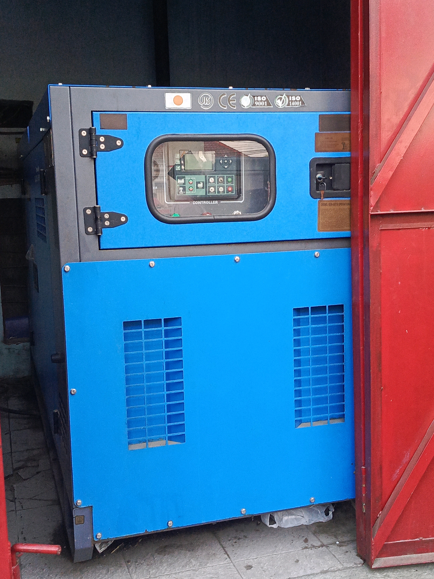 A blue and gray industrial generator is partially visible within a structure, with a red metal door partially open on the right side. The generator is labeled with various certifications including a Japanese flag, "JIS", "CE", "ISO 9001", and "ISO 14001". A control panel is visible behind a glass window on the upper section of the generator. The generator sits on a concrete surface, with a white plastic bag visible underneath its base. The environment appears to be an outdoor or semi-outdoor location, possibly a service area or industrial space. The location is identified as Gladak Anyar, Indonesia.
