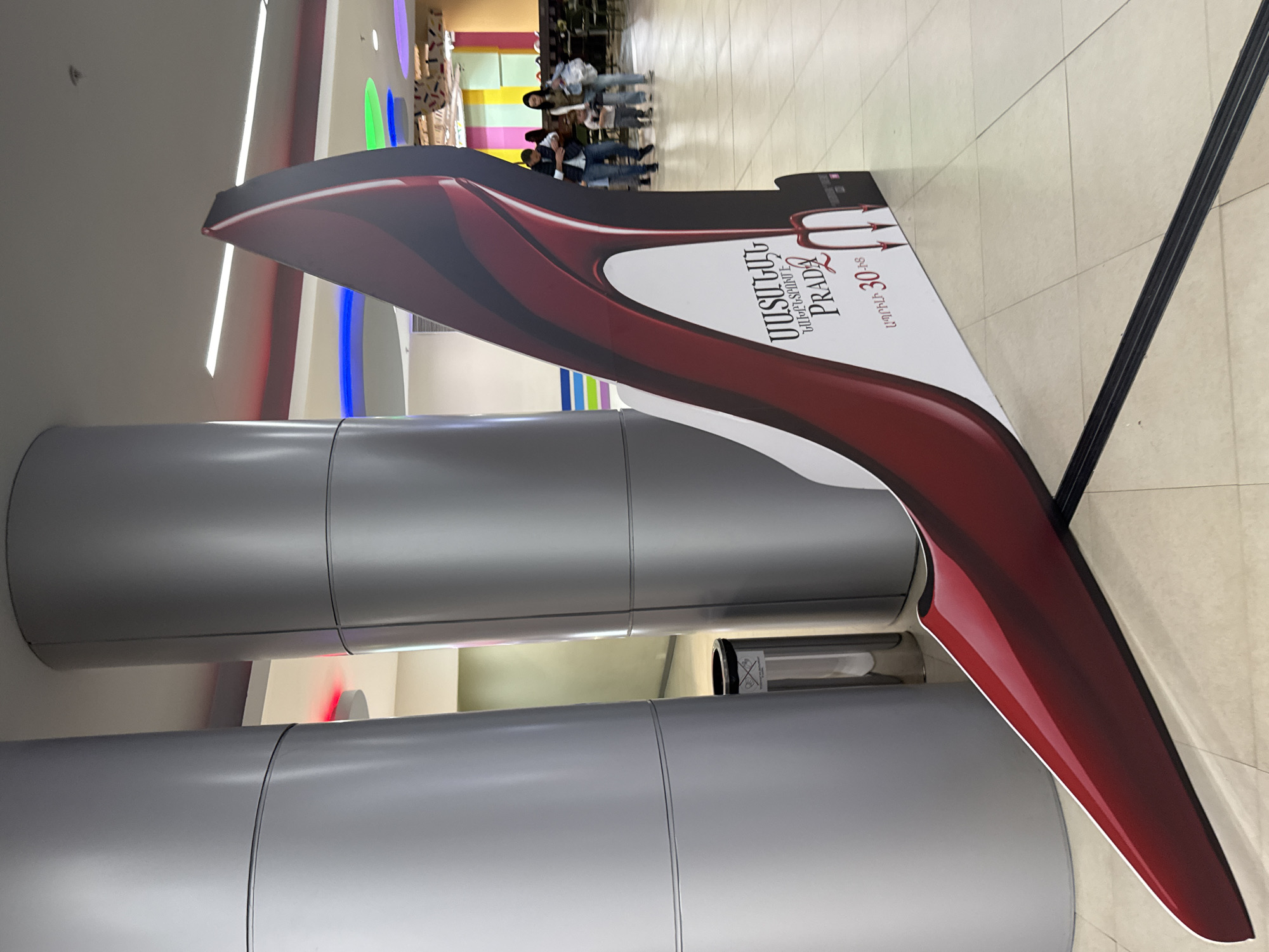 The image captures an indoor scene, likely within a shopping mall or large commercial building, featuring a prominent, large, stand-up advertisement shaped like a high-heeled shoe. The shoe display is predominantly dark red or burgundy with white accents, depicting a stylized design. Two large, silver-colored cylindrical pillars stand in the foreground, partially obscuring the view behind them.
In the mid-ground, behind the advertising display, several people are visible, appearing somewhat blurry. They seem to be walking or standing, though their specific activities are not clear. The floor is tiled with light-colored squares, and bright strip lighting is visible on the ceiling. A small black trash bin is discreetly placed between the two pillars.
Visible text on the shoe display is in Armenian and English. The Armenian text reads "ՀԱՍԱՐԱԿՈՒԹՅՈՒՆԻ ԹԱԳՈՒՀԻ PRADA" which translates to "THE QUEEN OF SOCIETY PRADA," with "GIRL OF SAINT" in smaller English text above "PRADA." Below this, "մինչև 30% զեղչ" is visible, meaning "up to 30% discount." The overall impression is an advertisement for Prada, likely a perfume or fashion item, in a commercial space in Yerevan, Armenia, as indicated by the Armenian text and provided context. The lighting suggests daytime indoors.