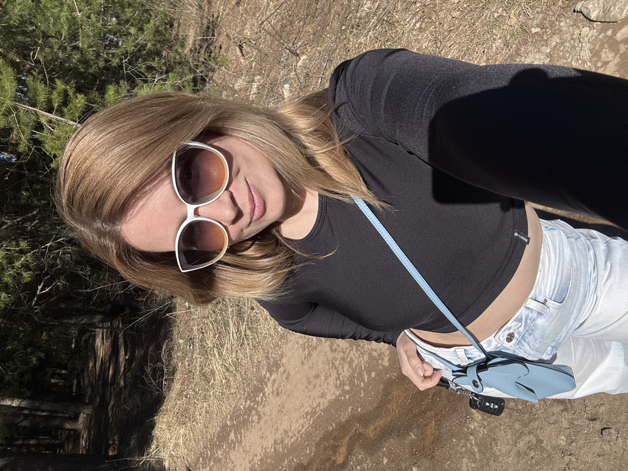 A woman wearing sunglasses, a black long-sleeved crop top, and white jeans is posing outdoors. She has a blue purse with a strap over her shoulder. The surroundings appear to be a natural setting, possibly a park or wooded area, with dry grass and trees visible in the background. The location is consistent with an outdoor area in Chaikovsky, Russia, characterized by natural landscape elements.