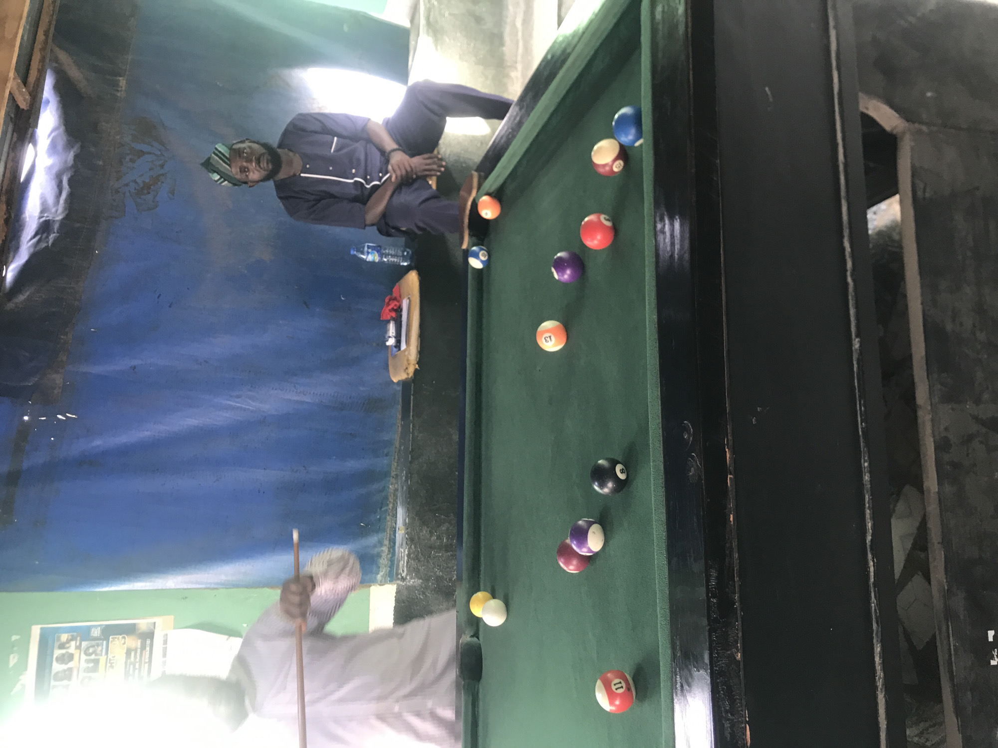 An indoor scene, likely a community space or game room, features two individuals around a pool table. On the left, a person, dressed in a light-colored long-sleeve shirt, is partially visible and in the process of striking a cue ball with a wooden pool cue. Their face is obscured by a bright light source. On the right, a second person is seated on a light-colored concrete ledge, observing the game. This individual is wearing a dark blue tunic and trousers with white trim, and a striped green and black cap. They have a beard and are looking towards the pool table.
The pool table, with a green felt surface and a black wooden frame, occupies the foreground and midground. Numerous pool balls are scattered across the table, including a white cue ball, a yellow 1-ball, a red 11-ball, a purple 4-ball, a black 8-ball, a red 3-ball, a yellow 13-ball, a purple 12-ball, a red 7-ball, a yellow/red striped 9-ball, and a blue 2-ball.
The background consists of a light green wall on the left, displaying a partially visible, brightly lit poster with images and text. The central and right background features a large blue tarp or fabric draped as a wall, with visible wrinkles and folds. A wooden beam runs across the top of this blue backdrop. Adjacent to the seated individual, a small, light-colored table or stool holds a clear plastic water bottle, a red cloth, and a small dark object. The floor beneath the table is dark and shows signs of wear. The overall lighting suggests natural light entering the space, with a strong glare on the left side of the image. The location is in Unknown, Unknown.