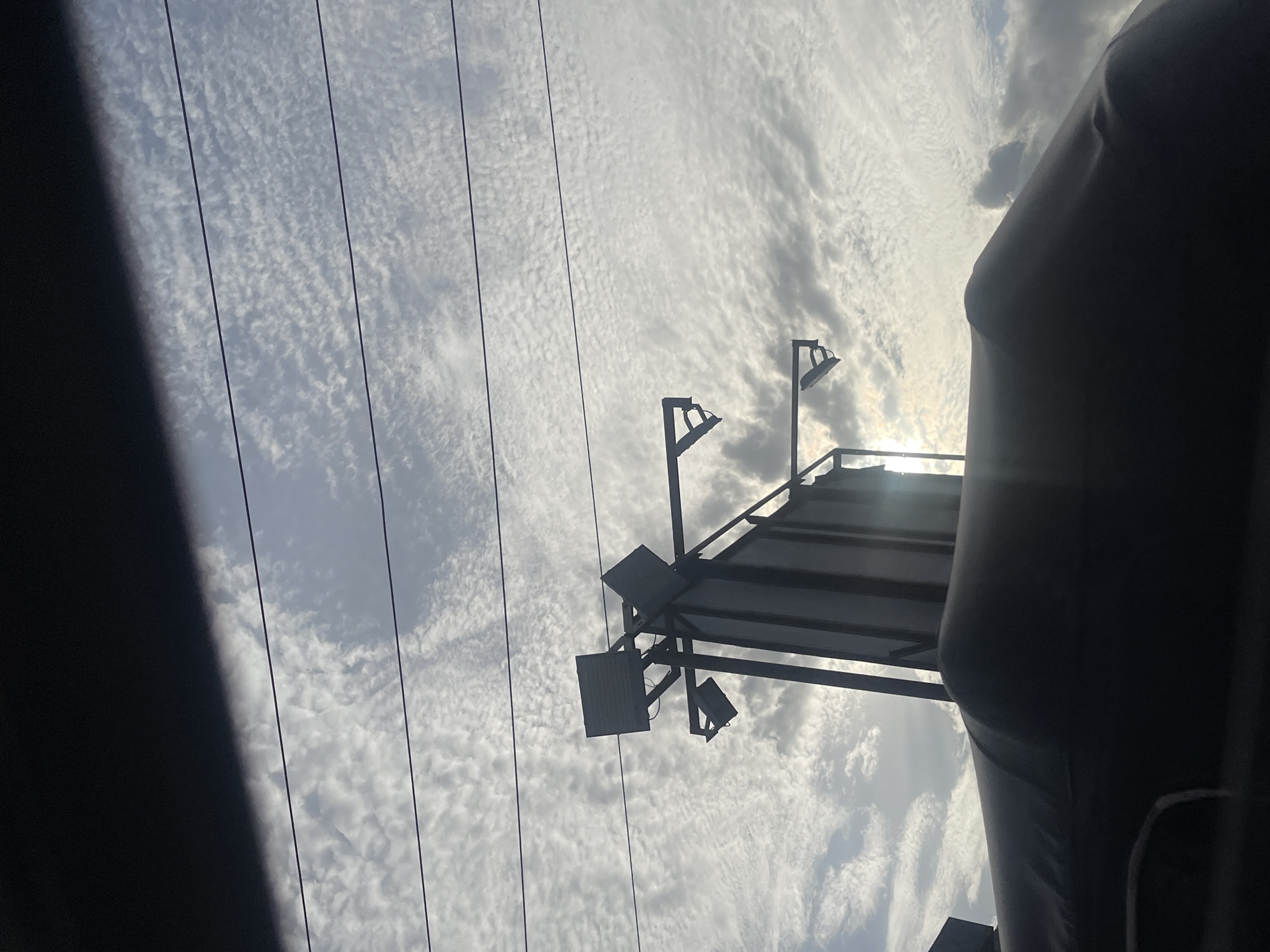 The image shows a cloudy sky in Ado, Nigeria, with power lines running across the view. A metal structure with lights is visible against the sky. Part of a large, dark, undefined object obscures the right side of the image.