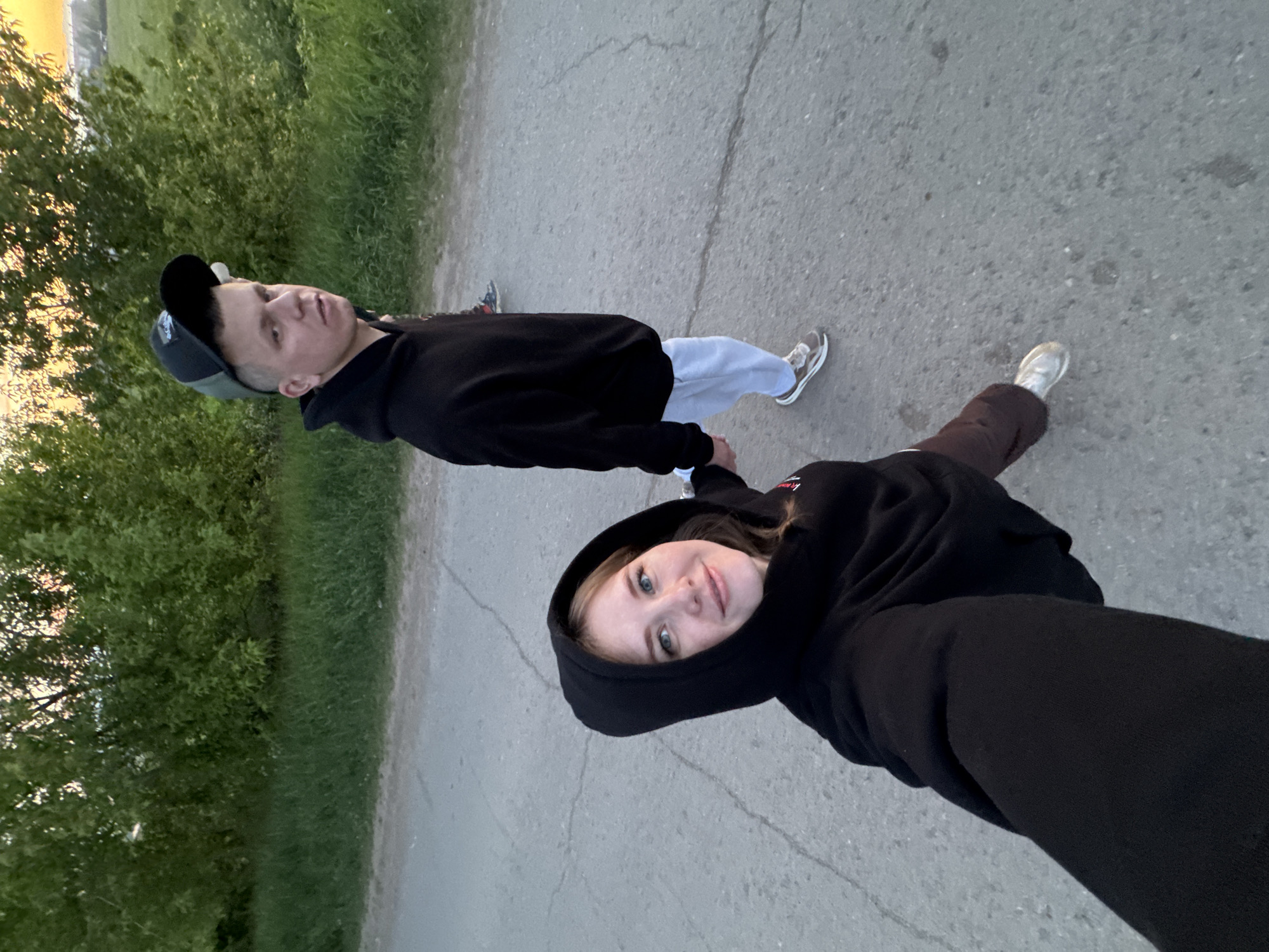 Two people, a man and a woman, are walking on a paved path. The woman is holding the camera, taking a selfie. They are both wearing black hooded sweatshirts. The man is also wearing a baseball cap and light-colored sweatpants, and the woman is wearing brown pants. They are holding hands. The location appears to be in Горный, Russia, with green foliage and grass visible to the left of the path. The lighting suggests it is dusk or early evening.