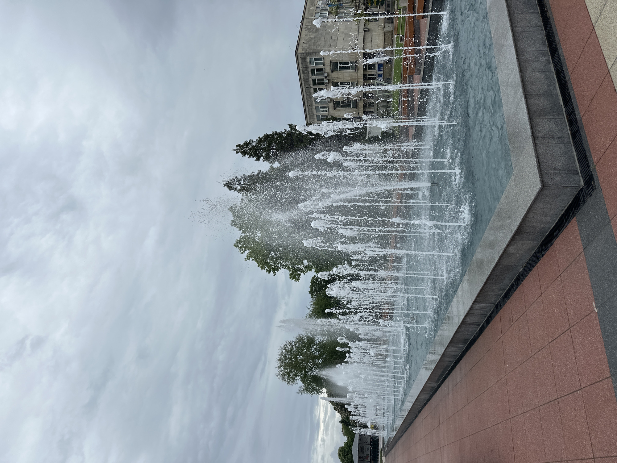The image captures a water fountain located in the city of Montana, Bulgaria. The fountain is rectangular in shape and features multiple jets of water shooting upwards, creating a dynamic display. In the background, trees and a building with visible windows can be seen. The ground surrounding the fountain is tiled with reddish-brown paving stones. The sky is overcast with cloudy conditions.