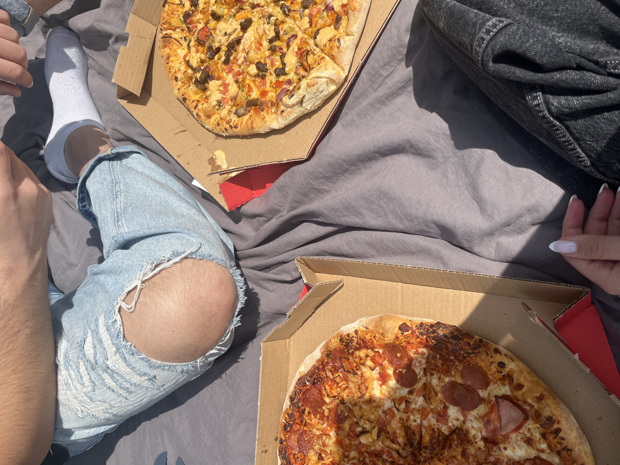 Two people are sitting on a gray surface with two pizza boxes. The person on the left is wearing ripped light blue jeans and a white sock. The person on the right is wearing black jeans. One pizza has pepperoni, and the other has various toppings. The location is not discernable.