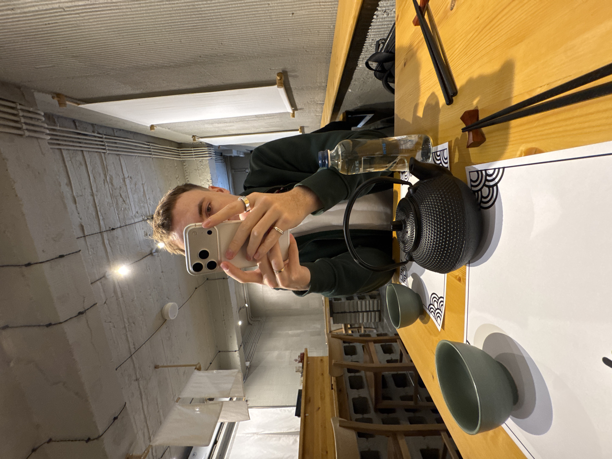 A person with short brown hair is taking a selfie at a table set for a tea ceremony in Tbilisi, Georgia. The person is wearing a dark green jacket over a white shirt. The table is made of light-colored wood and set with a black cast iron teapot, two small green teacups, and a bottle of water. Chopsticks rest on holders near the edge of the table. A white paper placemat with a decorative black and white pattern is placed in front of the tea set. In the background, chairs are visible, along with a gray concrete wall and ceiling with exposed pipes and ductwork. Hanging white lights are visible.