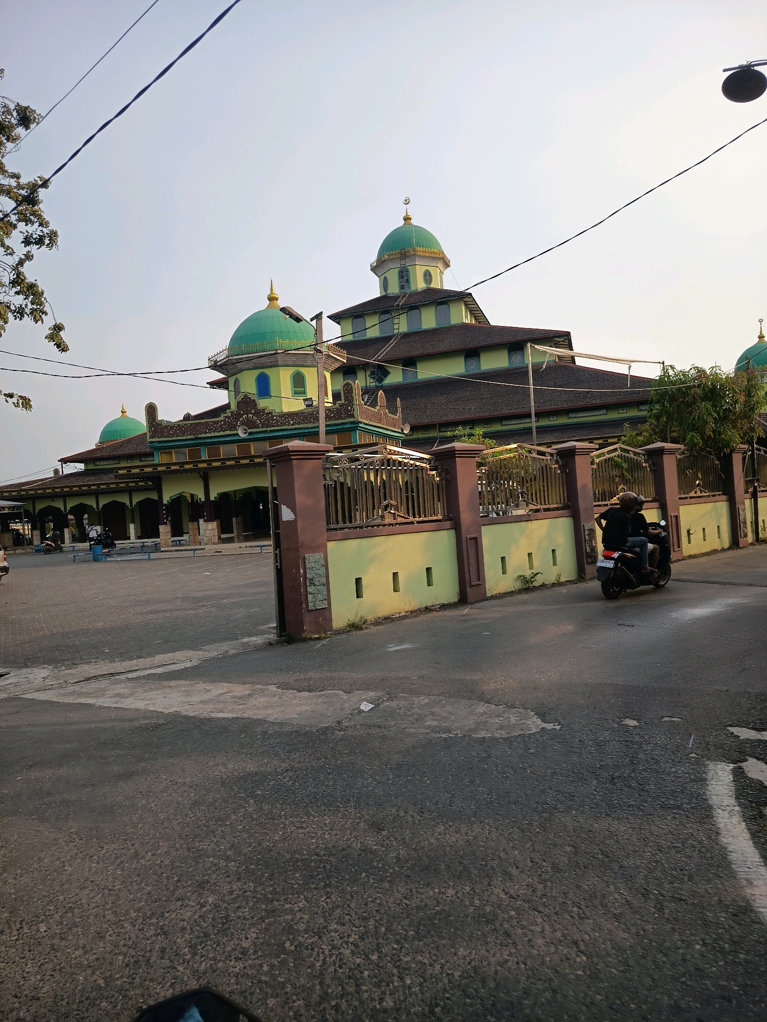 The image captures a street view in Banjarmasin, Indonesia, featuring a mosque with multiple green domes and a light green facade. The mosque's architecture is characterized by ornate details and arched entrances. A brown and light green fence with decorative elements separates the mosque's courtyard from the street. On the right side of the street, a motorcycle with two occupants, wearing helmets and dark clothing, is moving away from the camera. The street is paved with asphalt and shows some wear and tear. Trees are visible in the background, and power lines crisscross the sky. The sky is mostly clear with a pale, hazy quality. The overall scene depicts a typical urban environment in Indonesia with a prominent religious building.