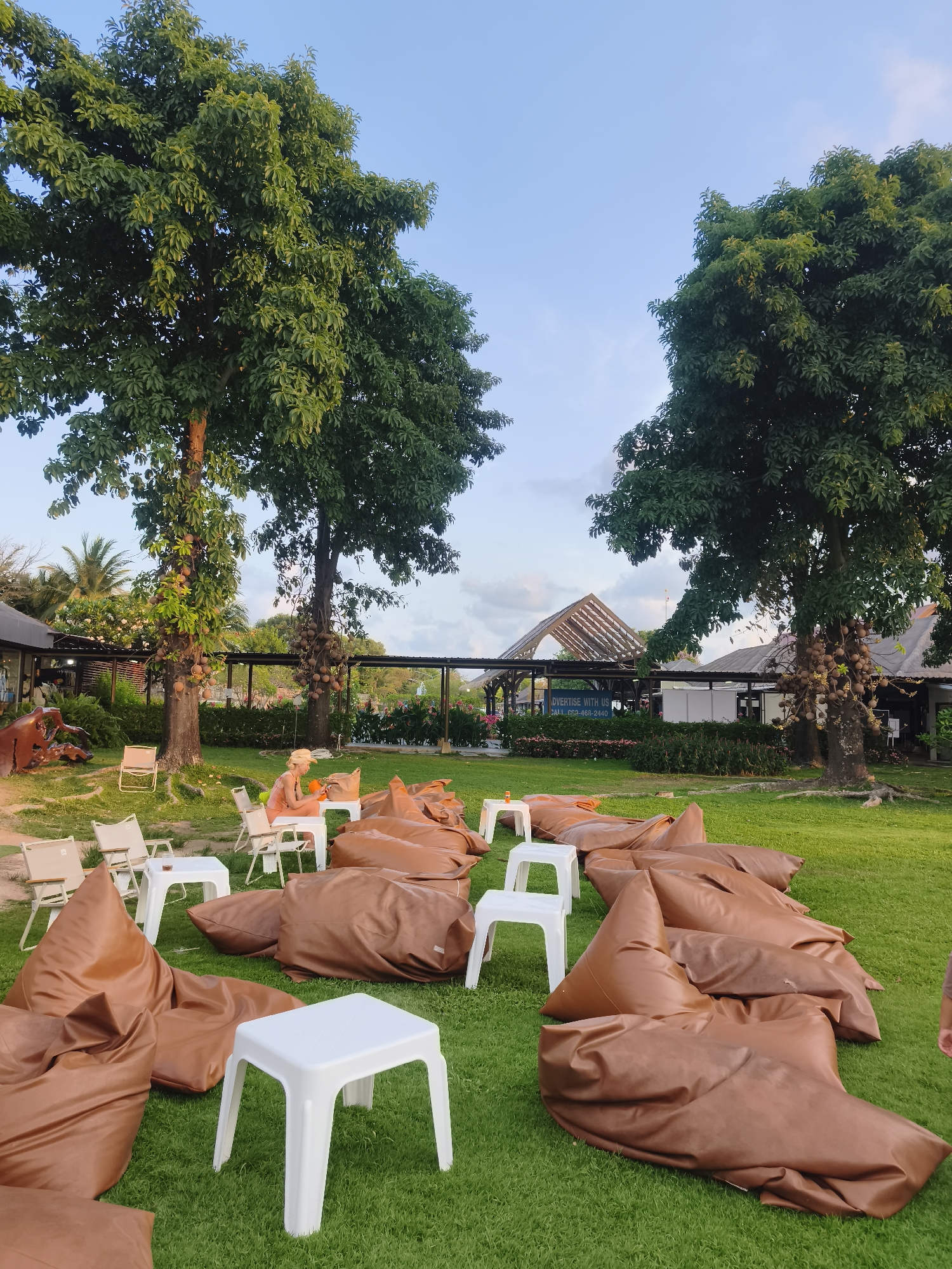 The image shows an outdoor lounging area on a grassy lawn in บ้านบางรักษ์, Thailand. Numerous brown beanbag chairs and small white plastic tables are scattered across the grass. Two large trees are positioned on the left and right sides of the frame. In the background, a woman wearing light orange clothing and a straw hat is seated at one of the tables. Beyond her, there is a structure with a partially visible sign that says "ADVERTISE WITH US CALL 662-468-2440". The sky is pale blue with some scattered clouds.