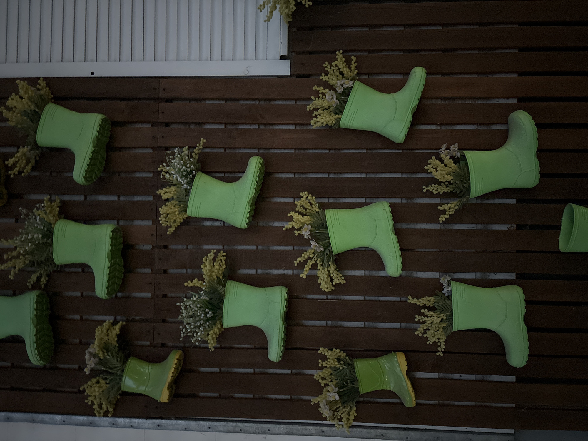 The image shows a display of green rubber boots hanging on a brown wooden slat wall. Each boot contains flowers. The display is located in the city of Chaikovsky, Russia. There are no people visible in the image. The environment appears to be indoors.