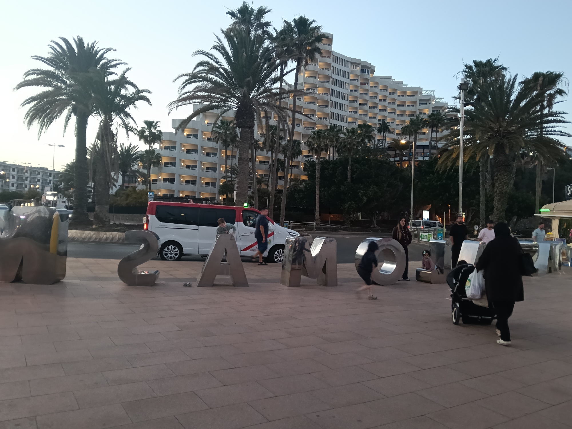 The photo depicts a scene at dusk in San Bartolomé de Tirajana, Spain, as indicated by the prominent "MASPALOMAS" sculpture. People are present, including a child running, a woman pushing a stroller, and other individuals standing or walking nearby. A white van with red accents is parked on the street behind the sculpture. The background features a multi-story white building with balconies and palm trees. The ground is paved with reddish-brown tiles. The sky is a pale color, consistent with twilight.
