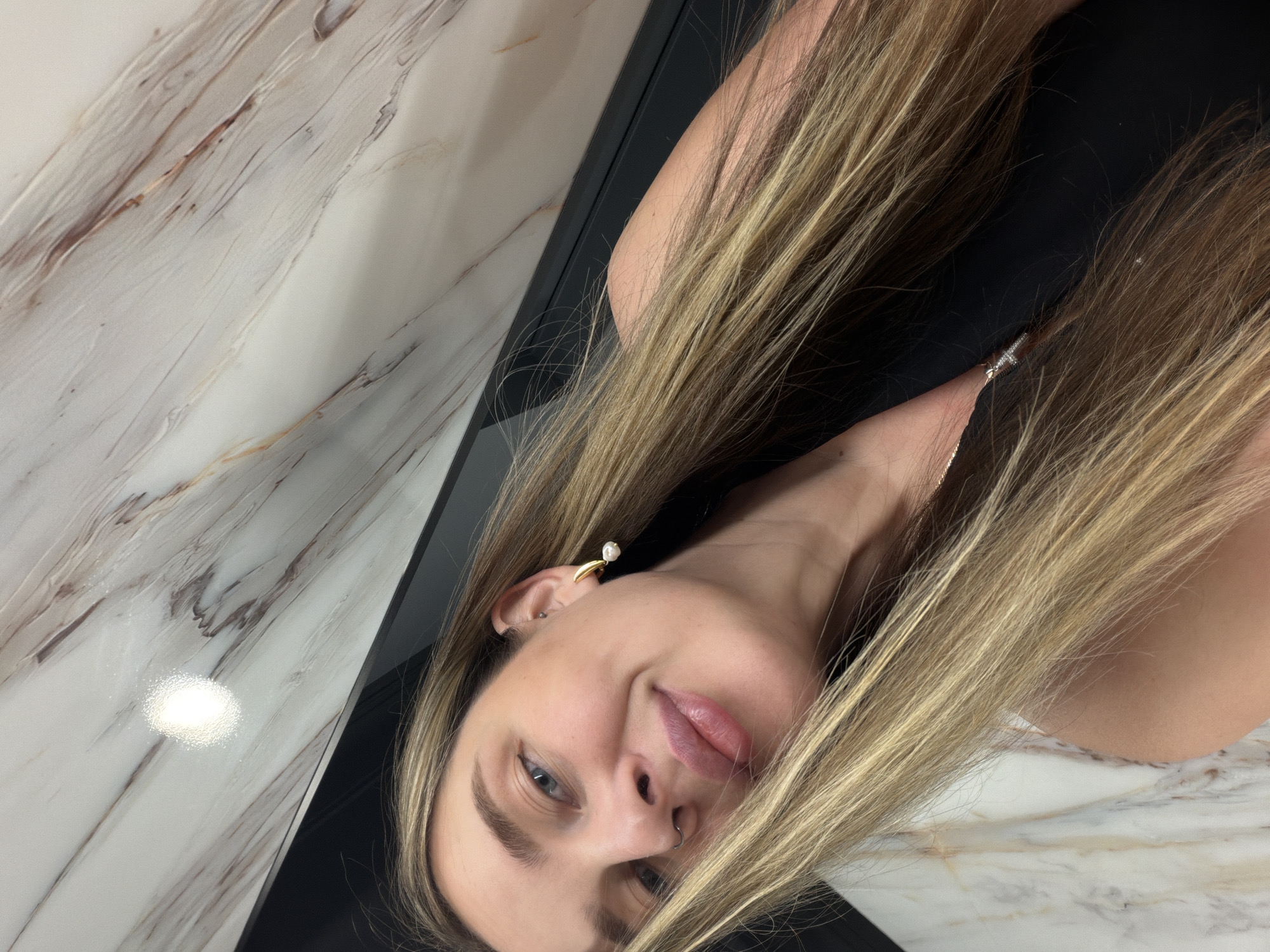 A fair-skinned woman with long blonde hair is seen close-up. She wears a gold earring with a pearl. The background consists of a marble-like wall and a black surface. There is no information available to determine the location is Moscow, Russia.