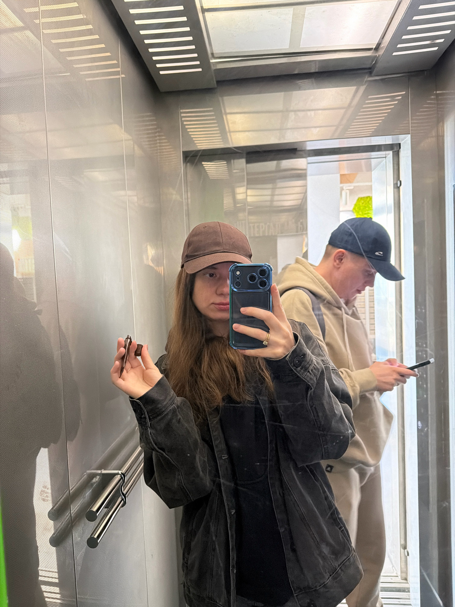 The image shows a young woman taking a selfie in an elevator. She is wearing a brown baseball cap, a dark denim jacket, and a black t-shirt. She is holding a small brown object, possibly a pen or a piece of candy, in her right hand. A man is visible in the background, also wearing a baseball cap and a light-colored hoodie, looking at his phone. The elevator has mirrored walls and metal handrails. The scene appears to be in an urban environment, possibly a residential building or a commercial space, with reflections of lights and possibly windows visible in the elevator walls. The text visible on the wall in the reflection suggests the location is Kyiv, Ukraine.