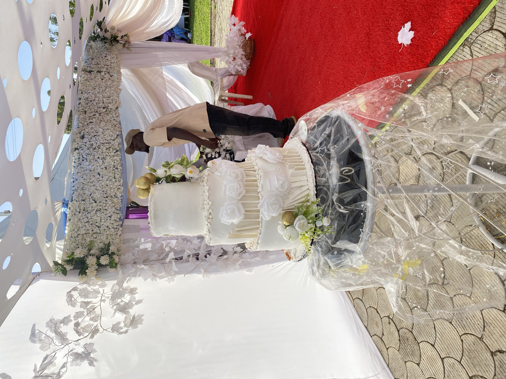 A three-tiered white wedding cake decorated with white roses and gold accents sits on a table covered in a clear plastic protector outdoors, under a white floral-decorated canopy. A man in a tan jacket and baseball cap stands next to the cake, seemingly preparing to cut it. A red carpet is visible in the background. The location is likely Bukuru, Nigeria.