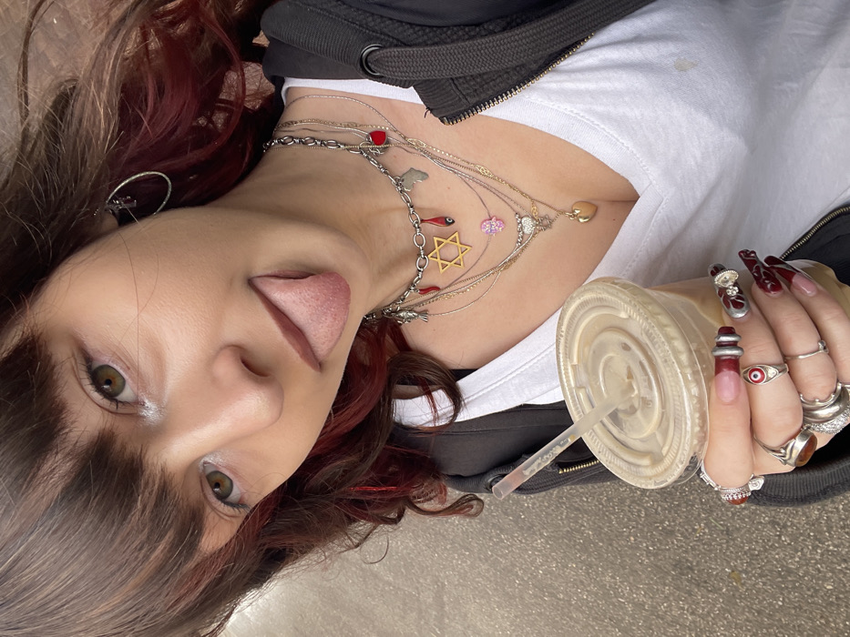 A young female subject is depicted from the upper torso upwards, with her head slightly tilted, looking directly at the camera. Her brown hair features reddish highlights and bangs. Her eyes are greenish-brown, and her tongue is partially extended from her mouth. She wears a dark-colored hooded jacket over a white V-neck t-shirt.
Multiple layered necklaces adorn her neck, including a prominent silver chain with various charms: a golden Star of David, a silver fish, a red heart, red chili or blood drop-shaped pendants, an evil eye, and a charm resembling the continent of Africa. Additional thinner chains feature a pink Hamsa hand and a small golden heart pendant. A silver hoop earring is visible in her left ear.
Her right hand, holding a clear plastic cup filled with an iced, light-brown beverage and a straw, is visible. This hand displays multiple silver-toned rings, including one with a red evil eye symbol and another with an amber-colored stone. Her fingernails are long, featuring a French tip style with intricate dark red designs and metallic accents.
The background is out of focus, revealing a textured, dark gray surface consistent with concrete or asphalt, indicating an outdoor urban environment in the city of Raanana, Israel. The scene is illuminated by diffused light.