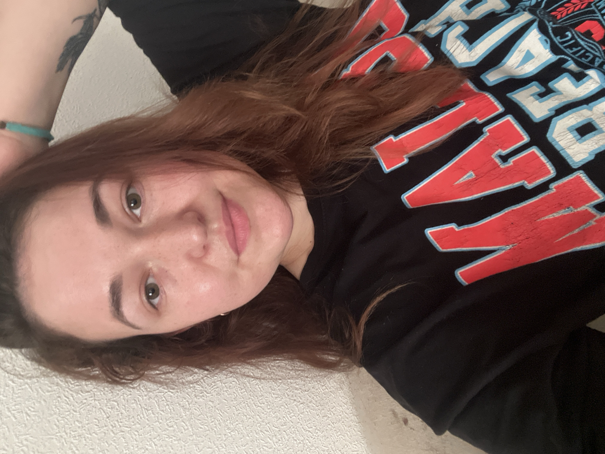 This is a vertical, eye-level, medium shot of a young woman with auburn hair, lying down and looking at the camera. She is wearing a black t-shirt with large red and blue lettering. The t-shirt appears to have distressed lettering. She has a tattoo on her left arm, partially visible, which appears to be a bird in flight. She is wearing a light blue bracelet on her left wrist. The background is a textured off-white wall. The lighting suggests it is indoors, possibly in the evening. The woman's expression is neutral or slightly smiling. The overall mood of the image is casual and relaxed. Visible text on the shirt includes "PACIFIC", and partial letters like "NATION", "OASIS", and "EST". The image was taken in Chelyabinsk, Russia.