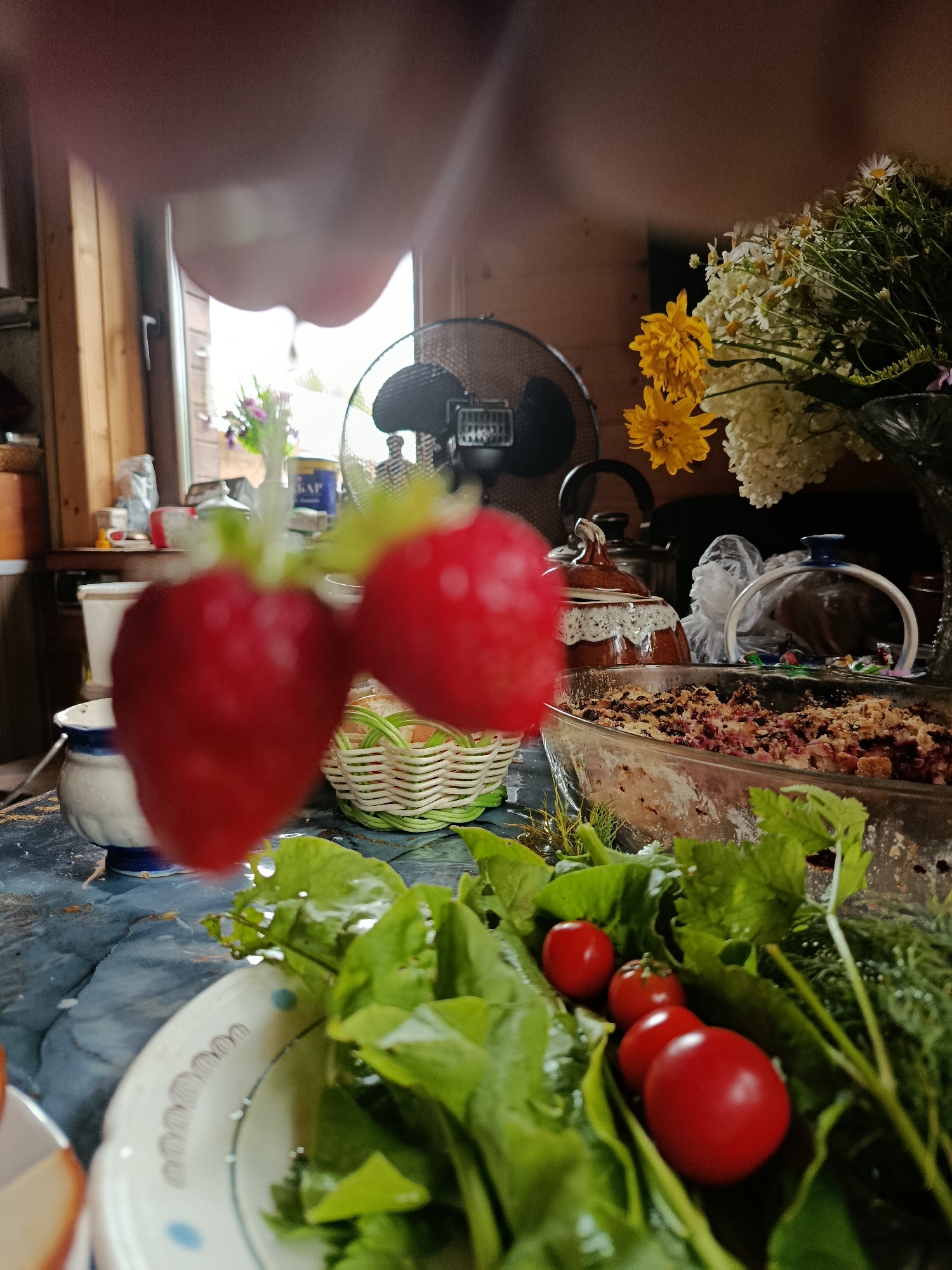 The image depicts a still life arrangement on a table, possibly in a rural setting in Vereya, Russia, given the hints. In the foreground, two out-of-focus strawberries are prominently displayed. To the right, a clear glass baking dish contains a baked dessert, likely a fruit crumble, with a topping of crumbled pastry and fruit. Surrounding the dish are fresh greens and several small red tomatoes.
In the middle ground, a small woven basket, a white and blue ceramic pot, and a teapot with a brown and white patterned lid are visible. Behind these items, a black oscillating fan stands on the table. Further in the background, a bouquet of flowers, primarily white daisies and yellow blooms, is arranged in a vase. The setting appears to be indoors, with wooden walls and a window providing natural light. The overall impression is a rustic, domestic scene.