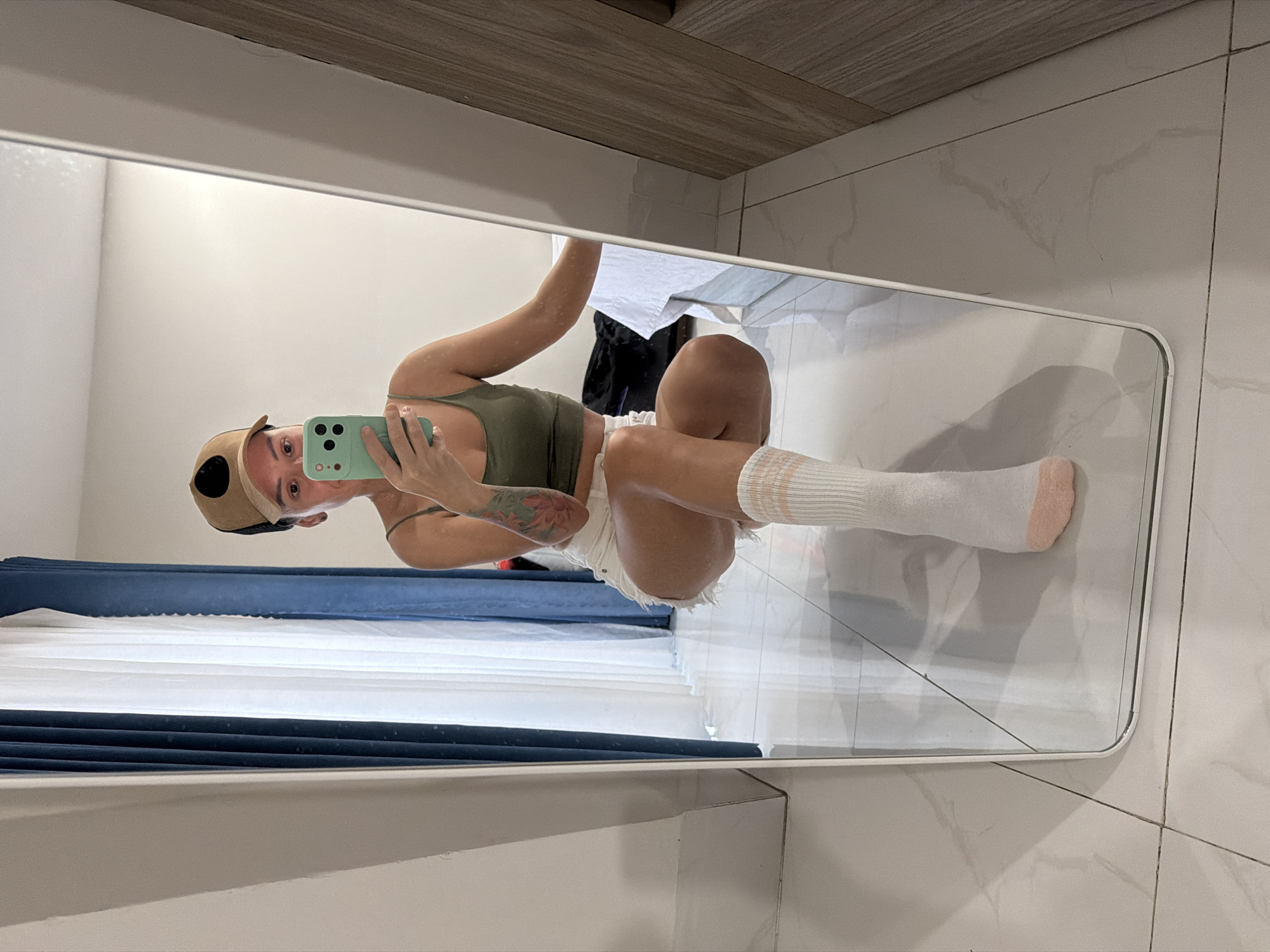 This is a mirror selfie taken by a person indoors, likely in a home or hotel room in Nha Trang, Vietnam. The person is wearing a green tank top, white shorts, a tan baseball cap, and white socks with pink stripes. They are holding a light green smartphone in their right hand, taking the photo. A tattoo of a pink flower with green leaves is visible on their left arm. The background reflected in the mirror shows white and blue curtains, a tiled floor, and a wooden cabinet. The lighting suggests it is daytime. The overall mood appears casual and relaxed. There is no visible text in the image.