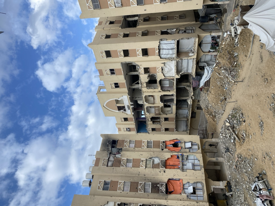This media file depicts a scene of severe urban devastation in Khan Yunis, Palestinian Territory. The image is dominated by multi-story residential buildings, likely apartment blocks, which have sustained extensive structural damage.
The central building features numerous large sections of its outer walls and internal partitions blown out, creating cavernous openings that expose the skeletal framework and interiors of multiple rooms across various floors. Windows are either completely missing, shattered, or partially covered with makeshift materials. Visible rebar, cracked concrete, and debris hanging from damaged floors highlight the severity of the destruction. In some of these openings on the buildings, white and orange tarpaulins are affixed, possibly as temporary attempts to seal off spaces or provide rudimentary shelter.
The ground level around and between the buildings is entirely covered with a thick layer of rubble, pulverized concrete, dirt, and general debris, indicating a significant destructive event. Towards the right side of the frame, a large white tarpaulin is spread on the ground, amid the wreckage.
The sky is a clear blue with scattered white cumulus clouds, suggesting it is daytime with fair weather. No people are visible in the image, emphasizing the desolation and the profound impact of whatever event caused this level of destruction. The scene portrays the devastating aftermath of conflict or a major incident. No discernible text is visible.