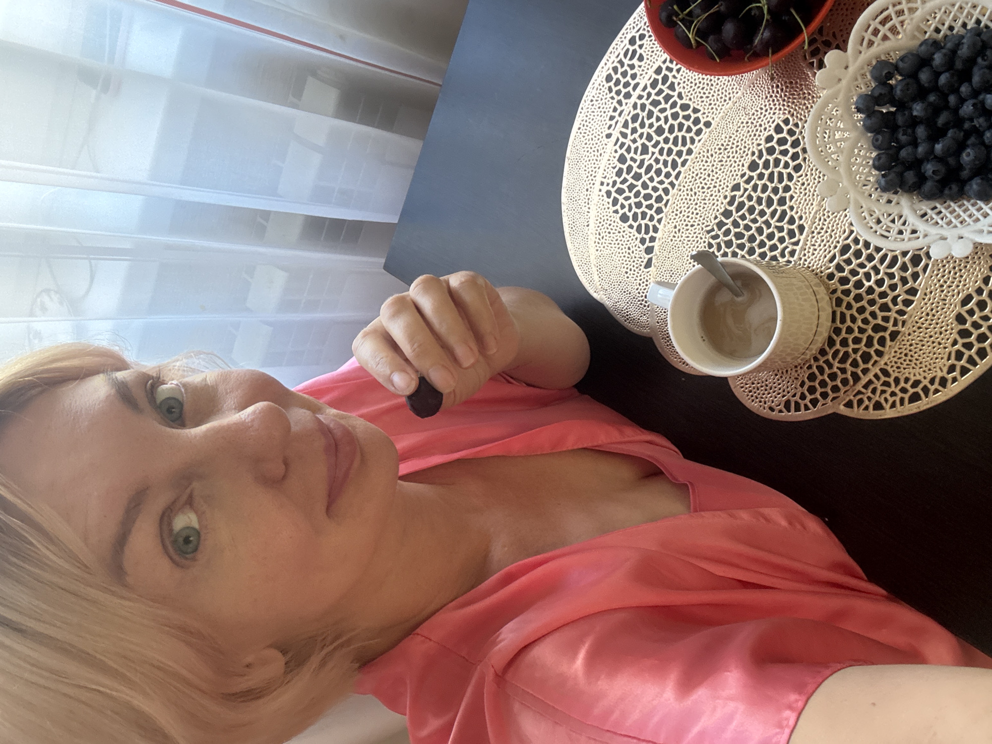 A fair-skinned woman with short blonde hair, wearing a pink satin robe, is seated at a dark table. She is holding a dark, round object, possibly a berry, between her fingers and bringing it to her mouth. In front of her on the table are a white mug with a spoon and a light brown beverage, a white woven basket filled with blueberries, and a red bowl containing cherries. The scene appears to be indoors, with sheer white curtains visible in the background, suggesting a residential setting. The lighting is bright, indicating daytime. The overall impression is a casual morning moment. The city of Sochi, Russia, is known for its coastal location and humid subtropical climate, which supports the growth of fruits like berries and cherries, making this a plausible setting for such a scene.