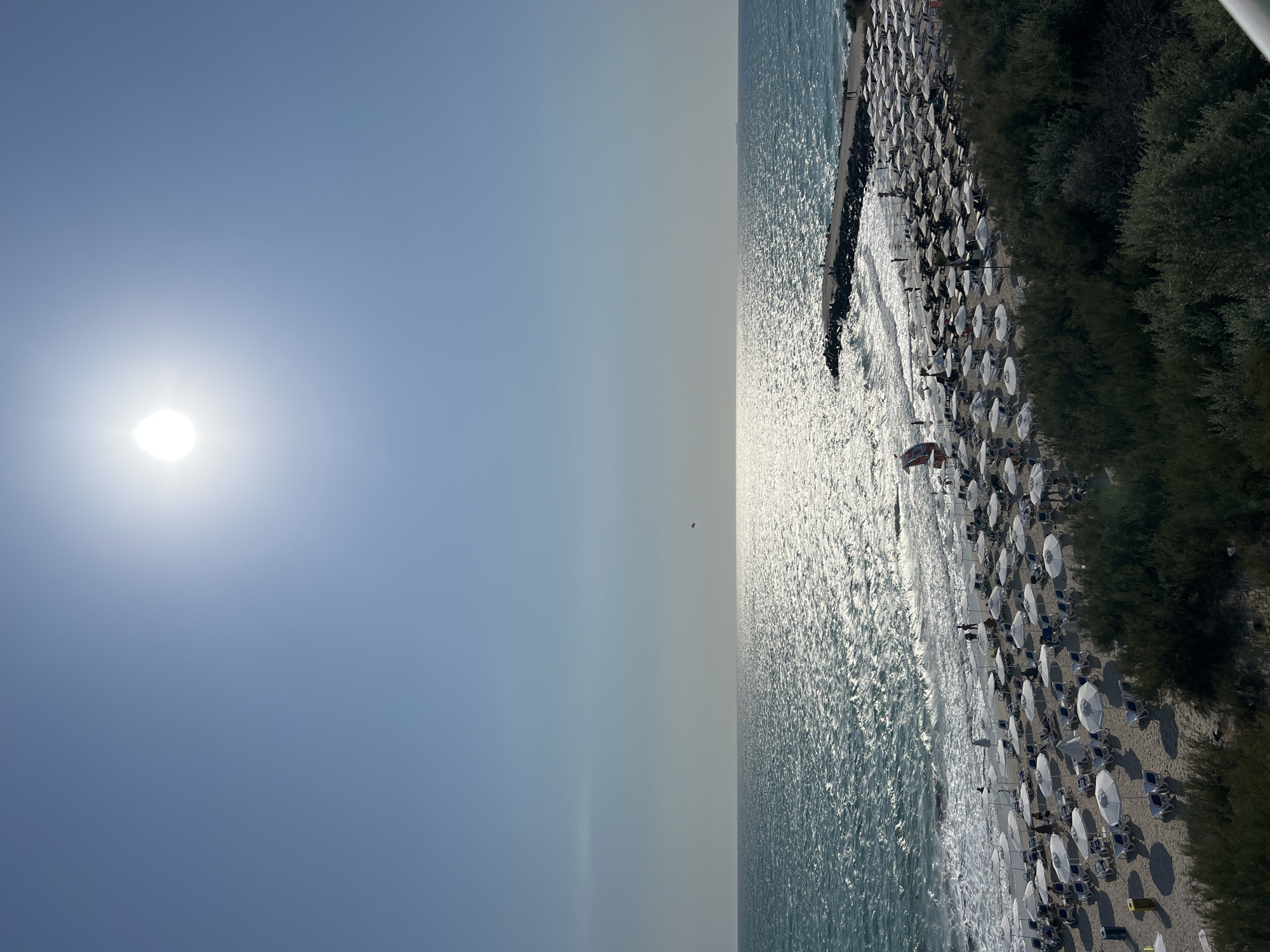 This is a high-angle, eye-level shot of a beach in Pomorie, Bulgaria, on a sunny day. The sun is high in the sky, casting a bright glare on the rippling, blue-green sea. A narrow, rocky pier juts out into the water on the right side of the frame. The beach is densely packed with white umbrellas and lounge chairs, with a few people visible walking or sitting. Green bushes line the edge of the sand. The scene conveys a typical summer day at a crowded beach resort.