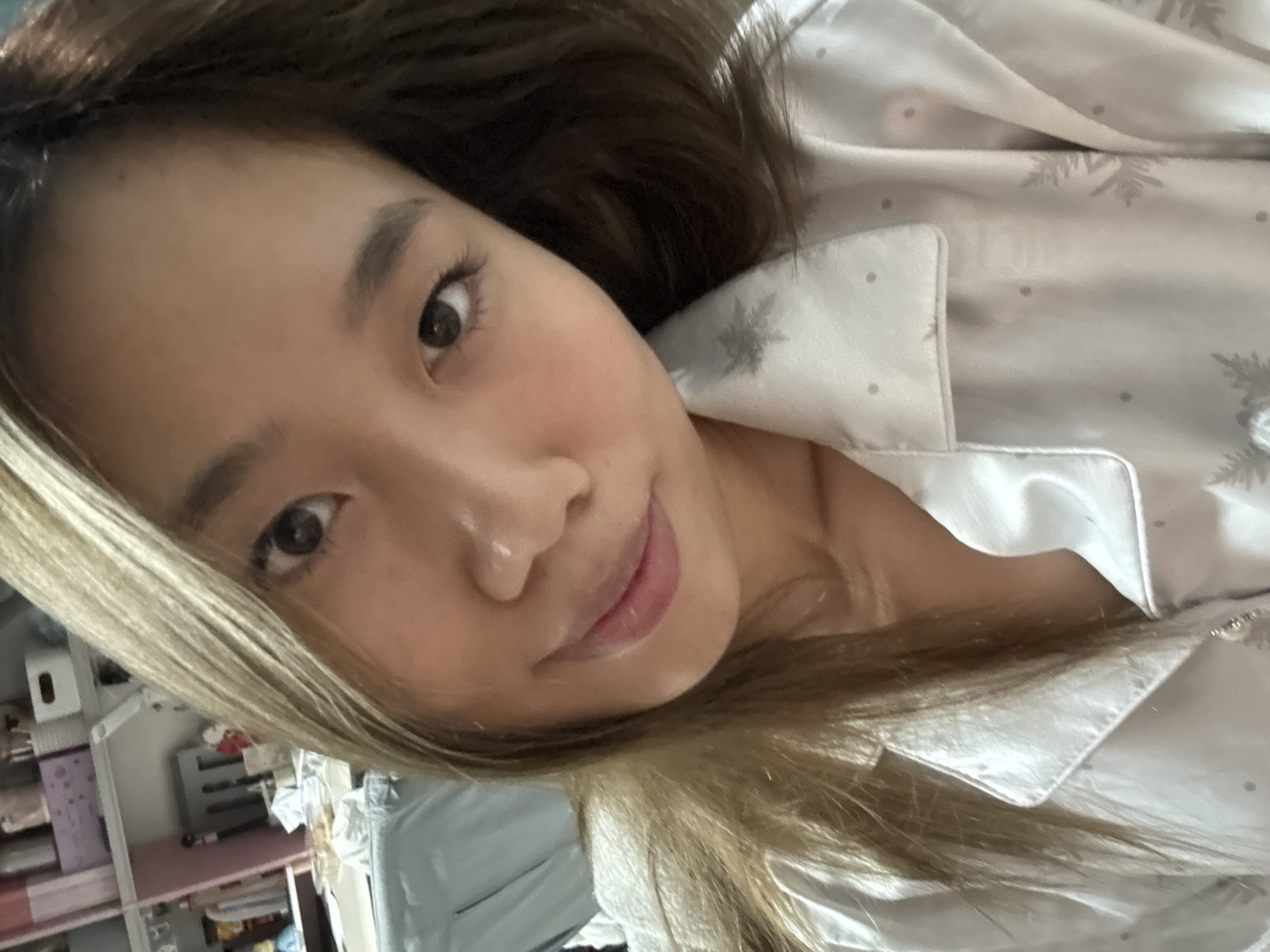 A young woman with light brown and blonde hair is angled towards the camera. She has brown eyes and fair skin. She is wearing a light-colored silky pajama top patterned with gray snowflakes and dots. The background is partially visible and appears to be a cluttered room with furniture, possibly a bedroom. There's no concrete evidence to establish that the picture was taken specifically in Nan'an District, China, nor are there sufficient details to confirm any actions or interactions, only the presence of the woman and objects in her environment.