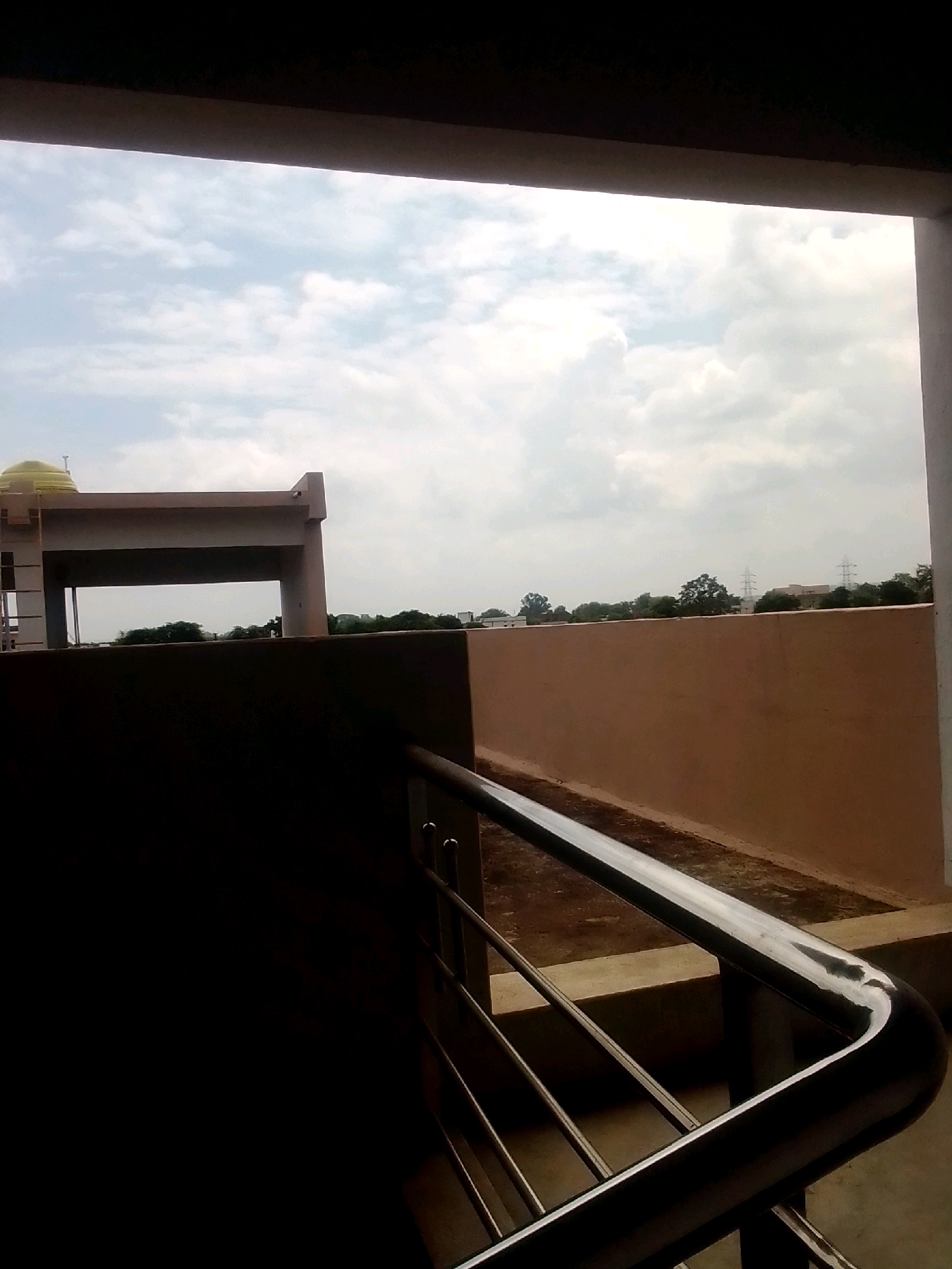 A close-up of a chrome railing in the foreground leads the eye toward a beige wall and a distant horizon. In the mid-ground, a pale yellow domed structure with a finial sits atop a modern building with a large rectangular opening. Beyond the building, a line of trees separates the structures from a cloudy sky. In the far distance, two sets of electrical towers are visible. The image is taken from a shaded area, possibly a balcony or walkway, looking out over an urban landscape.