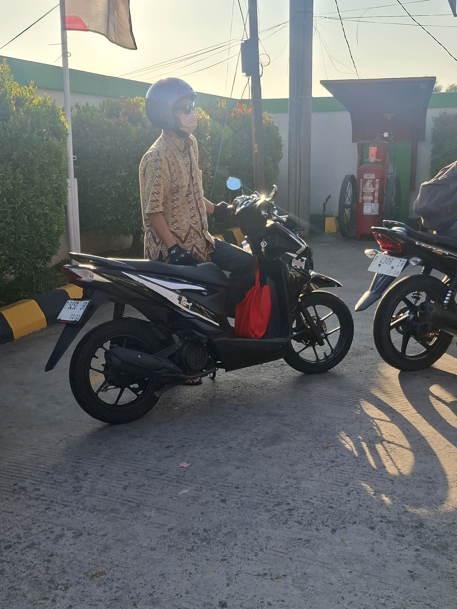A medium shot shows a person wearing a patterned shirt, a dark helmet, and a face mask, sitting on a black scooter with a red bag hanging from the handlebars. The scooter is parked on a paved surface. In the background, there is a row of green bushes, a flagpole with a flag, a tall pole with wires, and a red fire extinguisher on display. To the right of the scooter, another black motorcycle is partially visible. The scene appears to be set outdoors, possibly near a gas station given the presence of the fire extinguisher and what looks like a fuel pump structure behind the bushes. The lighting suggests it is late afternoon or early morning with long shadows cast on the ground. The license plate of the scooter reads "F 5450 UR". This scene is located in the city of Sukabumi, Indonesia.