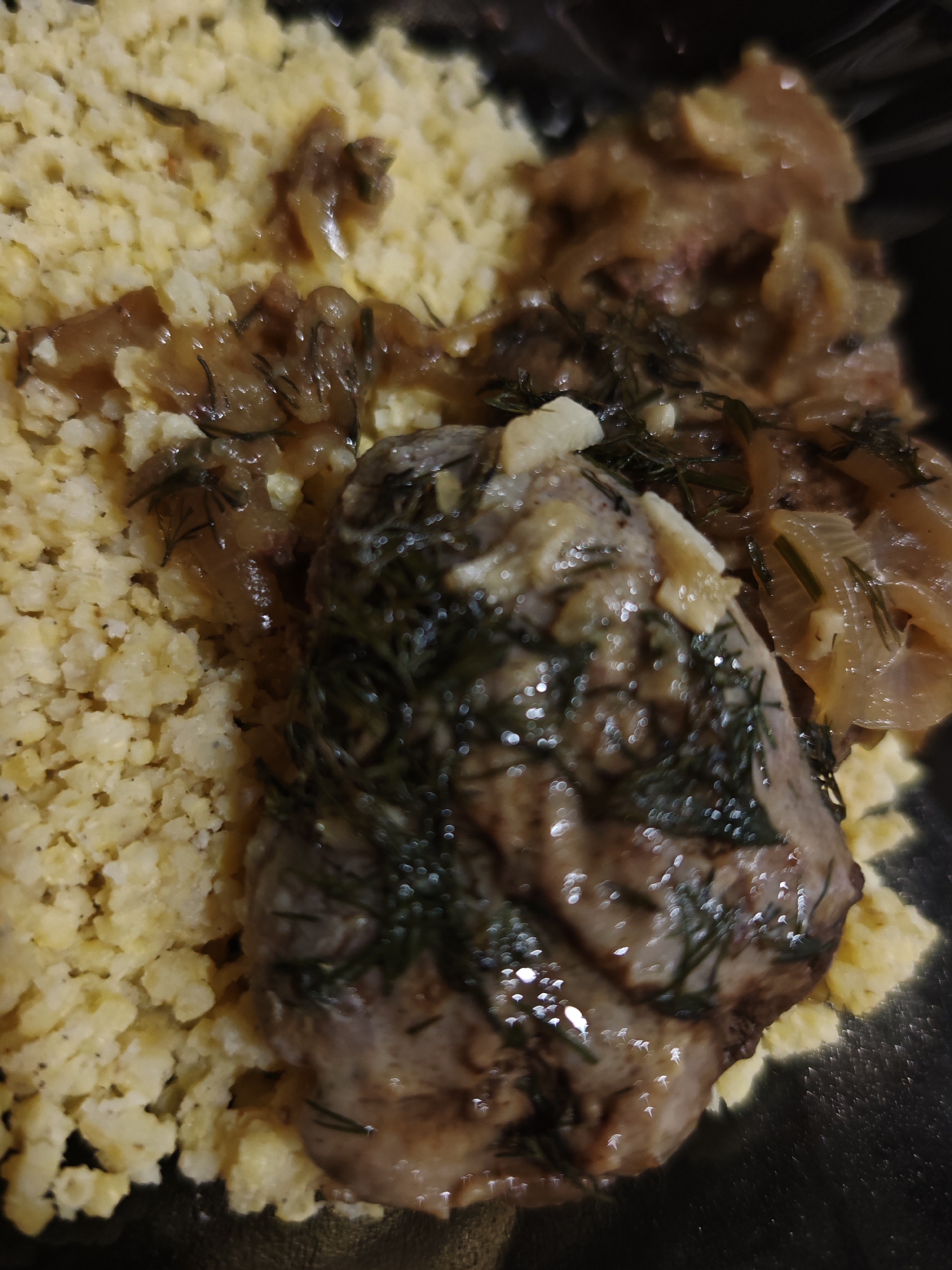A close-up, overhead shot shows a serving of food, possibly a meal in a dark container. The dish consists of a mound of bright yellow couscous on the left side, with a portion of meat, likely beef or pork, with a dark brown sauce and herbs, predominantly dill, placed on top of and next to the couscous. The meat appears to be braised or stewed, with visible pieces of onion mixed into the sauce. The lighting in the scene creates numerous specular highlights on the surface of the food, suggesting a glossy sauce and moist texture. The background is dark and out of focus, with the food occupying the majority of the frame. The location is listed as Unknown, Unknown.