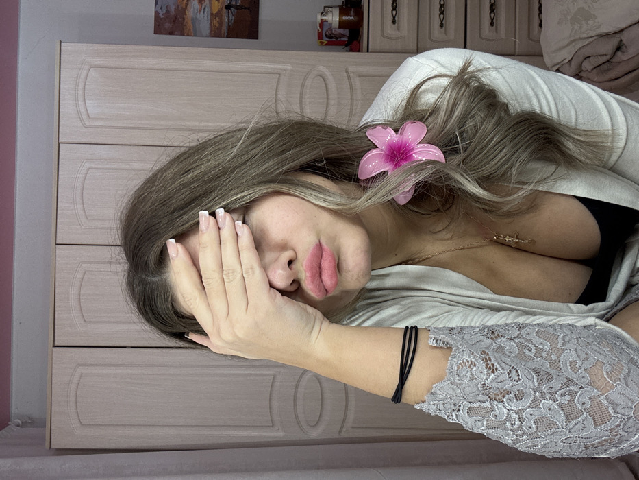 The image depicts a female subject in an indoor residential setting, consistent with a bedroom or dressing room, stated to be in Chaikovsky, Russia. The exact location within the city is indeterminable from the visual information provided.
The subject is a female with long, wavy, light brown or blonde hair, styled to her right. Her right hand is raised, covering her eyes with the palm facing inward and fingers slightly splayed; the fingernails display a white-tipped French manicure. Her lips are pursed, displaying a pink lipstick application. She is wearing a light-colored, possibly grey or beige, cardigan or robe with a deep V-neckline over a dark-colored garment, likely a bra or camisole, revealing cleavage. A gold-colored chain necklace with a crucifix pendant is visible around her neck. The left sleeve of her outer garment features grey lace detailing. Three thin black bands, likely hair ties, are worn on her left wrist. A translucent pink flower accessory is clipped into her hair on the right side of her head.
The background primarily features a light-colored wooden wardrobe with four panel doors, extending across the left and center. Above the wardrobe, a section of a pink-painted wall is visible. To the right, a dark-framed picture depicting a sunset or sunrise over water with a silhouette of a person on the shore is mounted on the wall. Below the picture, a light-colored dresser or nightstand with two drawers and metal handles is partially visible. On its surface, a red container, a white pump bottle, and other indistinct items are present. A light-colored, crumpled fabric, possibly bedding or a blanket, is seen in the bottom right corner.