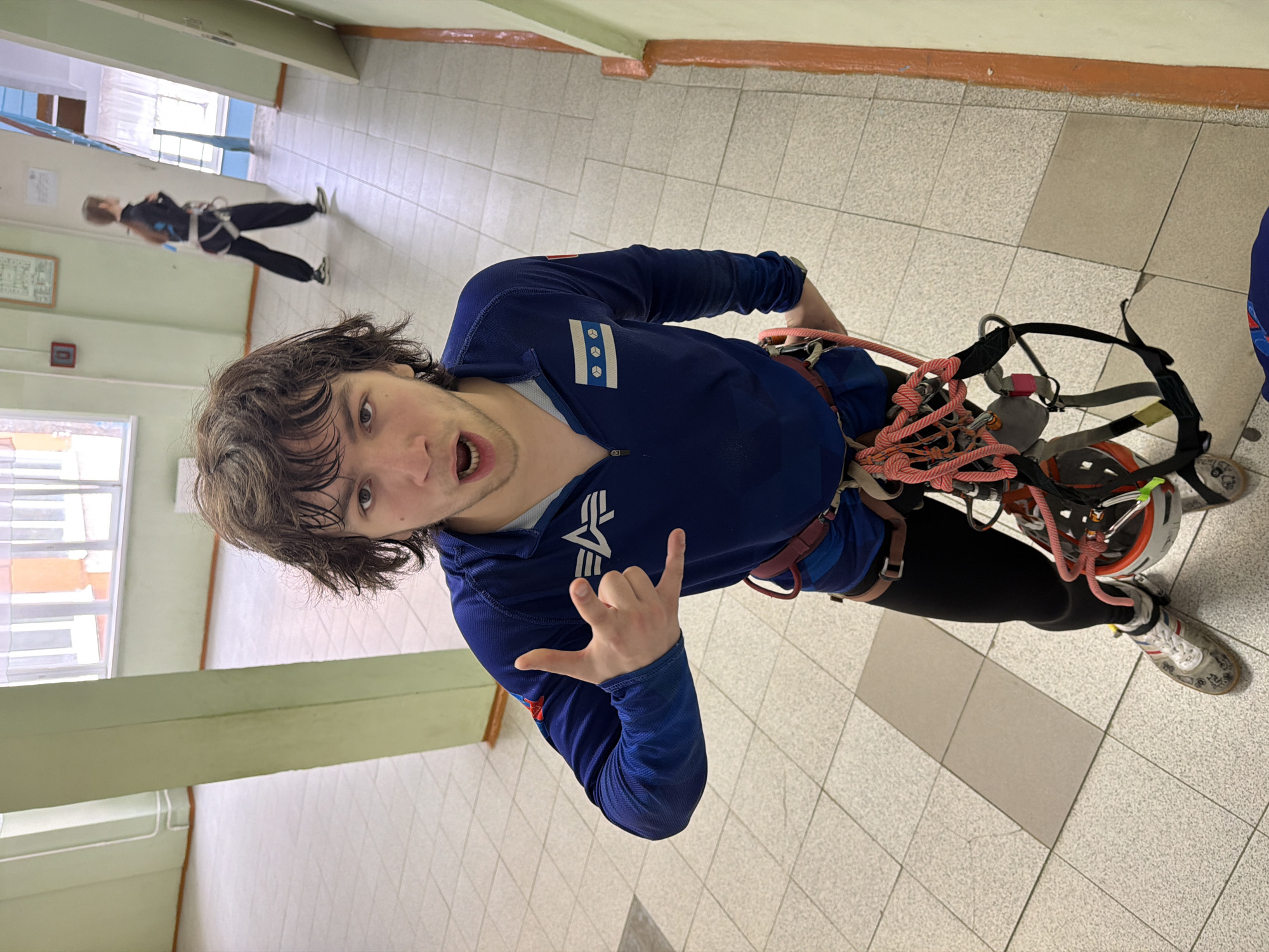 A young man wearing a blue long-sleeved shirt with the Alpha Industries logo and a Chicago flag patch is standing indoors, posing for a selfie with a rock climbing harness on. He is giving a "rock on" hand gesture. In the background, another person is walking down a hallway, also wearing a climbing harness. The location appears to be a building interior with tiled floors and painted walls. The city and specific location in Berezniki, Russia, cannot be identified from the image.