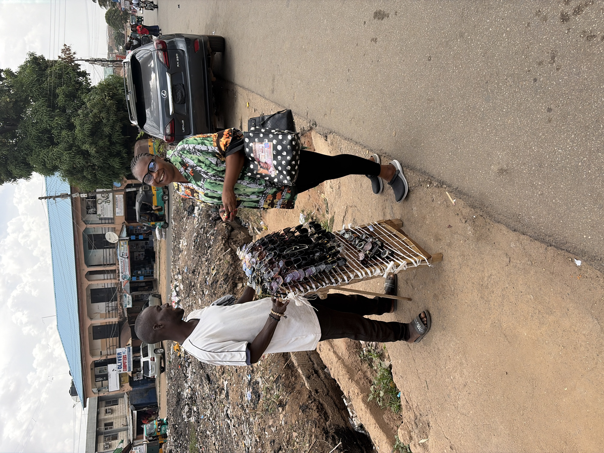 The image captures a vibrant street scene in Jos, Nigeria, focusing on an interaction between two individuals. In the foreground, a woman with glasses and a friendly smile, dressed in a colorful patterned shirt, black leggings, and sneakers, is looking towards a man. She carries two bags, one black with white polka dots and an image, and another plain black one.
The man, presumed to be a vendor, stands opposite her, holding a wooden display stand laden with numerous sunglasses. He wears a white short-sleeved shirt, dark trousers, and dark sandals, with beads on his wrist.
They are positioned on the paved roadside, next to a rough dirt shoulder filled with visible litter and debris. Behind them, a dark grey Lexus RX 350 SUV is parked. Further back, a row of buildings with corrugated metal roofs and various storefronts line the street. Visible signs include "OLIVER COMPUTERS LLC" and another partially visible sign reading "GODWIN MC...". Other vehicles, including a tricycle and motorcycles, can be seen further down the road, indicating a busy area. Utility poles and power lines crisscross the background. The sky is overcast, suggesting daytime with diffused light. The overall atmosphere appears lively, typical of an urban street in Nigeria, with the woman's smile indicating a pleasant interaction, possibly involving the purchase of sunglasses.