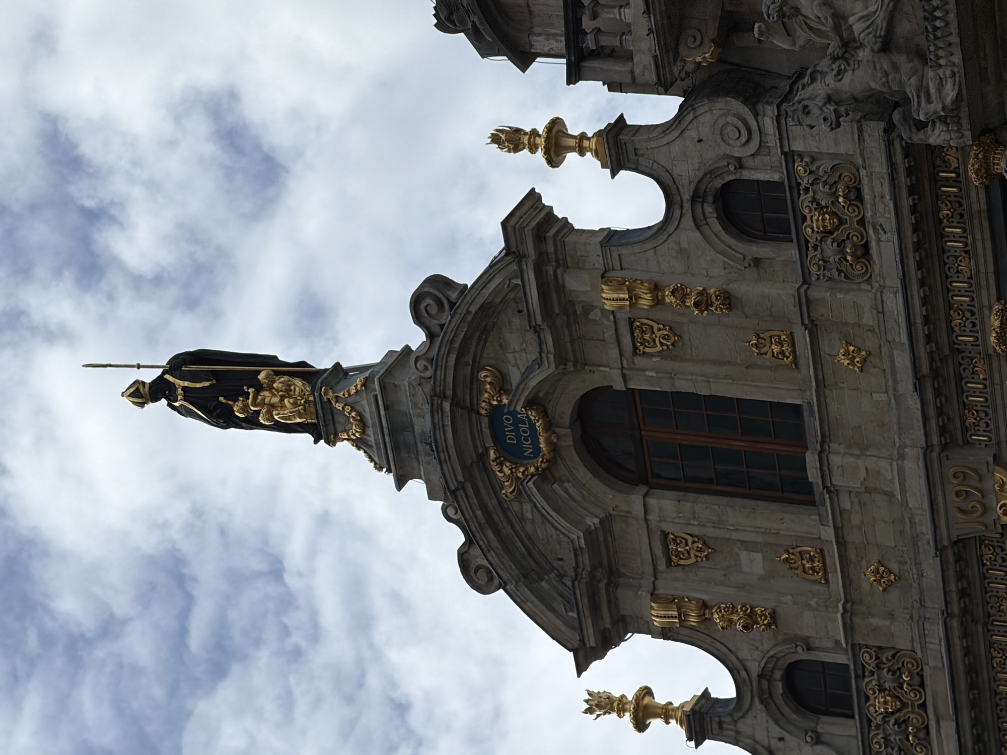 The image shows the Guildhall of the Brewers (Maison des Brasseurs) in Brussels, Belgium, with its statue of St. Arnould of Soissons, the patron saint of brewers, on the top, against a cloudy sky. The architecture is ornate with detailed stonework and gold ornamentation. The facade features windows, gold adornments, and the inscription "DIVO NICOLAO". The building showcases rich historical architectural styles common in the Grand Place of Brussels.