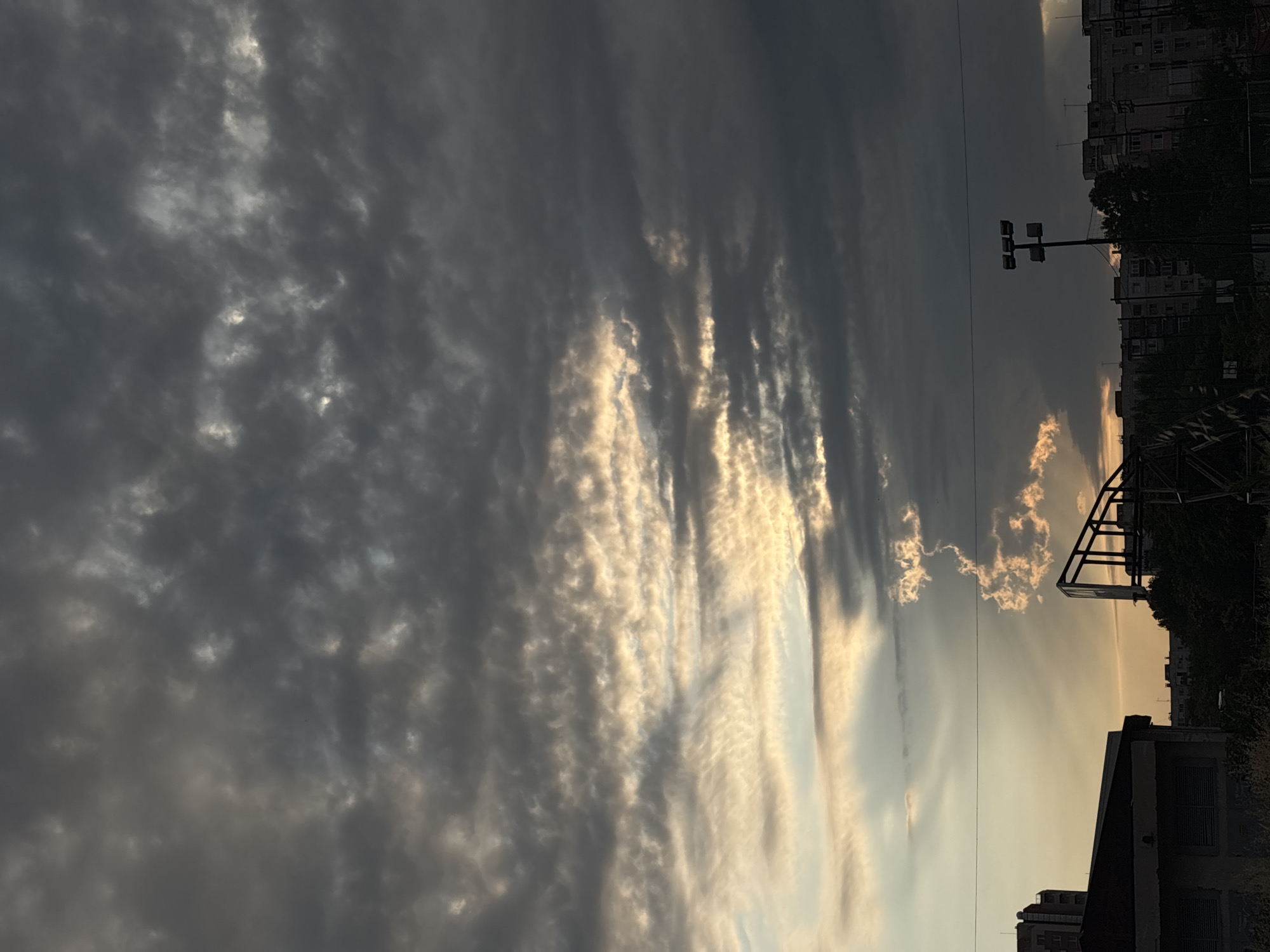 The image depicts a cloudy sky with the sun breaking through in patches, casting golden light. The scene appears to be an urban setting, likely in the city of Unknown, Unknown. In the foreground, there is a dark, angular structure, possibly part of a stadium or industrial building, with some vegetation growing around it. Further in the background, residential buildings and a sports floodlight are visible against the dramatic sky. The overall impression is one of a potentially overcast or stormy day with a break in the clouds.