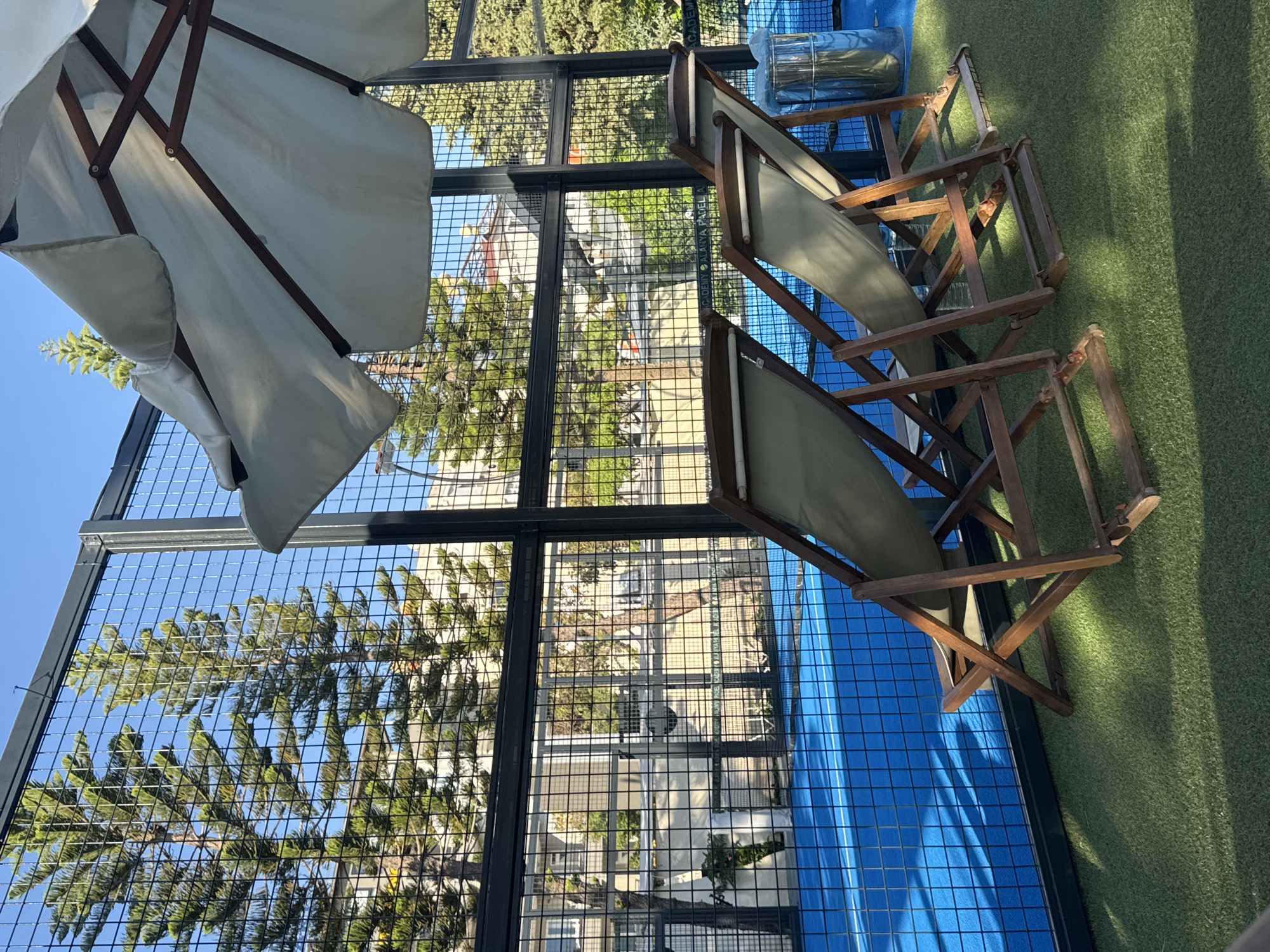 The image captures an outdoor scene in Alanya, Turkey. The perspective is from a high vantage point, looking down onto a balcony or patio enclosed by a black metal wire mesh fence. Partially visible in the upper left corner is a white patio umbrella with a wooden frame. Two folding wooden beach chairs are positioned on the balcony. One chair is on a green artificial turf area, while the other is partly on the turf and partly on a blue surface (likely a painted concrete or a blue covering).
Through the fence, there is a view of the surrounding urban environment, including buildings with red roofs and lush green trees. The outdoor space shows glimpses of other buildings. The overall impression is a relaxed, semi-private outdoor space with elements of a recreational area. The lighting indicates it is daytime, likely mid-day, with shadows cast by the furniture and umbrella.