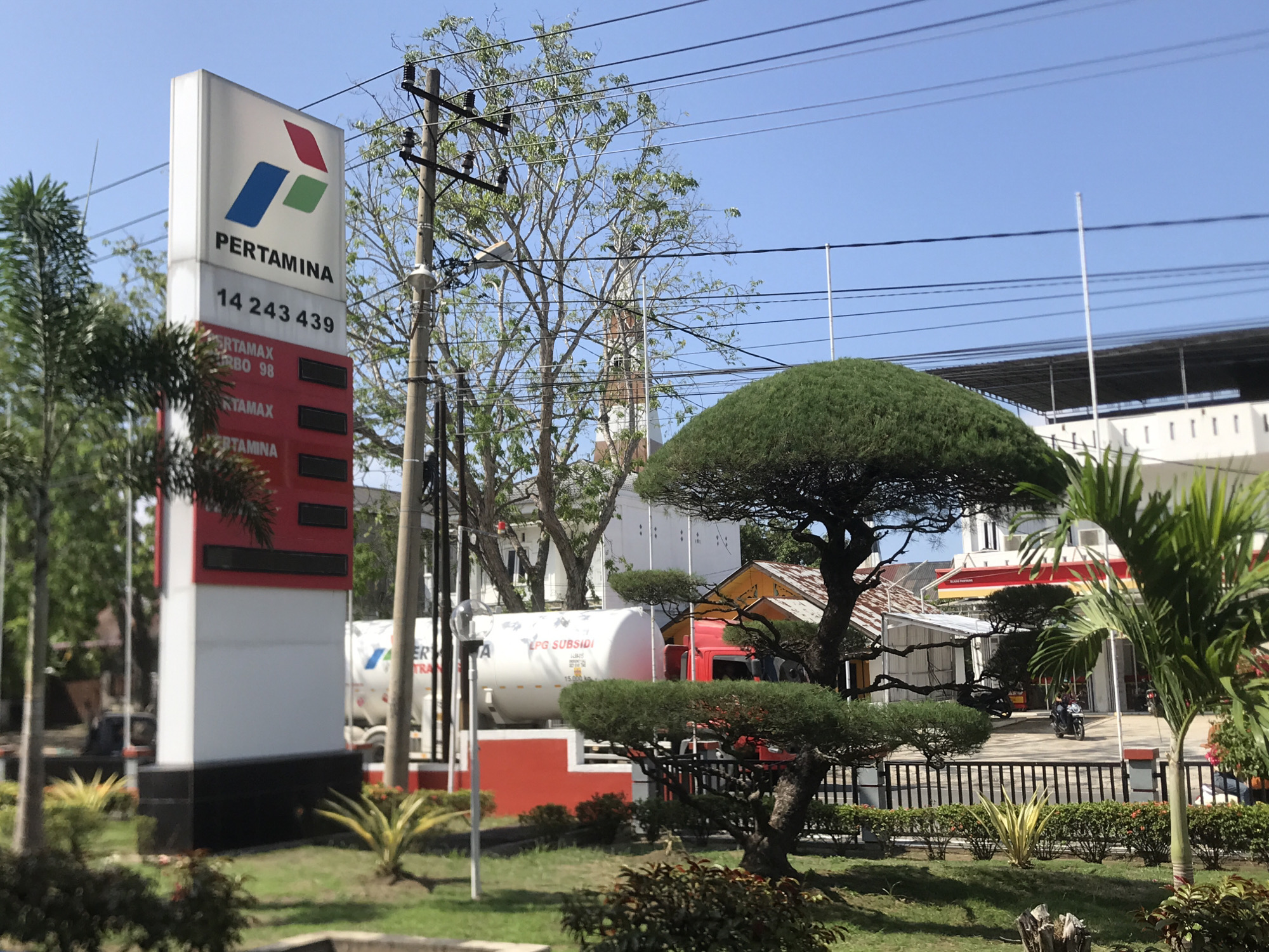 Here is a detailed description of the image:
The image shows a Pertamina gas station in Lhokseumawe, Indonesia, on a sunny day. A tall, white sign with the Pertamina logo and pricing information stands prominently on the left. The sign displays the name "PERTAMINA" and the phone number "14 243 439." The prices for various grades of gasoline, including "PERTAMAX TURBO 98" and "PERTAMAX," are listed.
Behind the sign are two large white tanker trucks with the words "LPG SUBSIDI" printed on them. The gas station has a red curb and is landscaped with palm trees and other greenery. A unique, umbrella-shaped tree is visible on the right side of the image. A black iron fence separates the landscaping from the station itself.
In the background, a building with a white facade and a red awning is partially visible. Power lines run across the top of the image. A utility pole stands near the Pertamina sign.
A few people are on motorcycles in the background, indicating activity at the gas station.