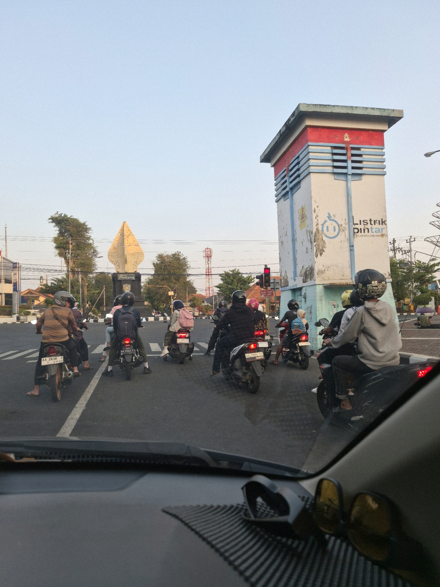 A street scene in Purworejo, Indonesia, features a traffic intersection with multiple motorcycles and riders. On the right side of the image is a tall, narrow, white tower with red and blue decorative accents, bearing the text "Listrik Pintar" and a logo resembling a stylized electrical plug. In the background, a golden, leaf-shaped monument stands on a dark pedestal, surrounded by trees and a telecommunications tower. The sky is a pale blue, suggesting either early morning or late afternoon. The perspective is from inside a vehicle, with the dashboard and a pair of sunglasses visible in the foreground.