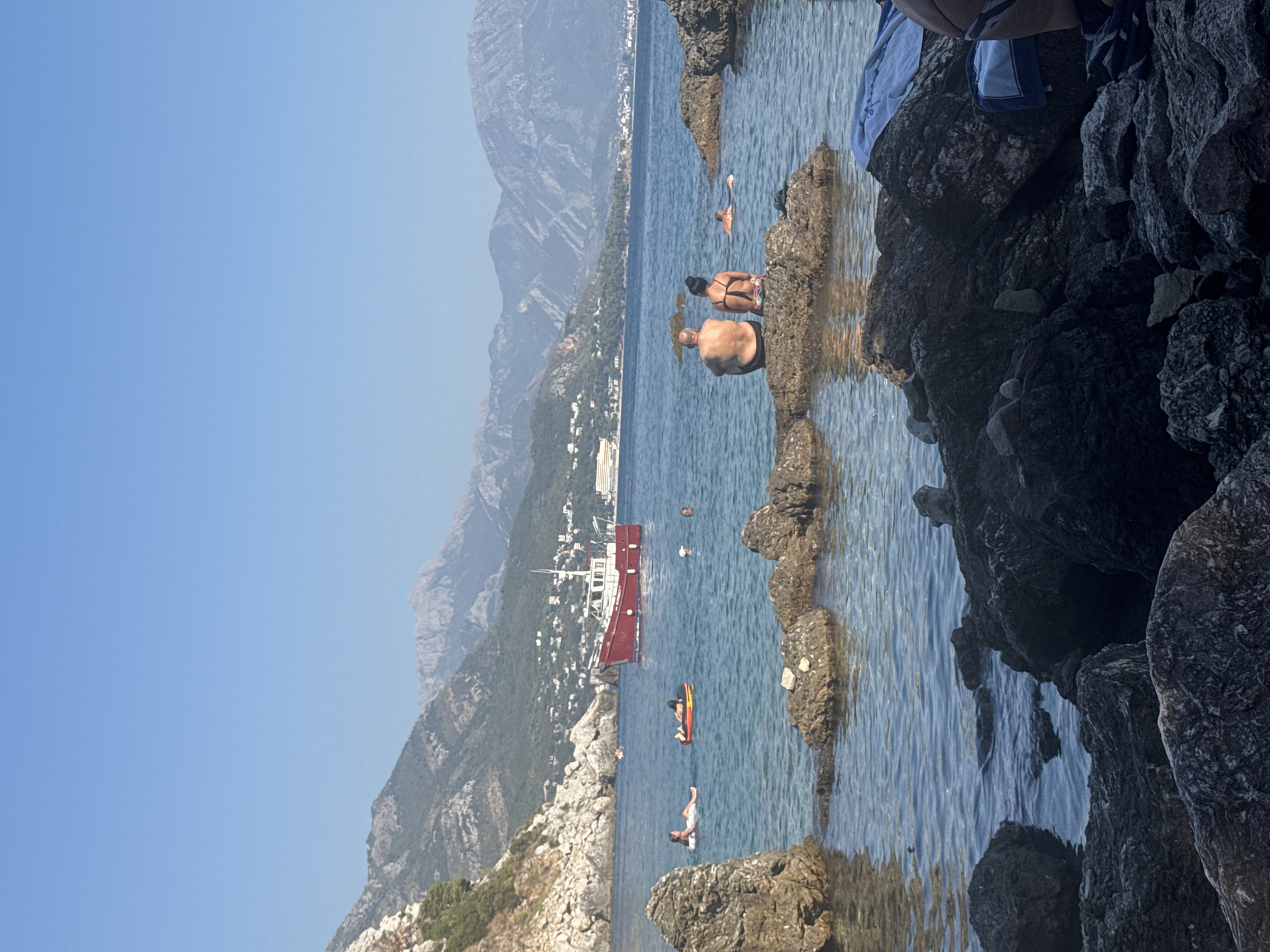 The image depicts a rocky shoreline on a clear, sunny day in Sutomore, Montenegro. The sea is a calm, transparent blue, with several people enjoying the water. In the foreground, large, dark rocks form a natural barrier, with smaller rocks submerged in the shallows. A couple, viewed from behind, is seated on one of these rocks, facing the sea. Further out in the water, three individuals are visible, two on inflatable floats and one swimming. A red and white boat is sailing in the middle ground. The background features a mountainous coastline, dotted with buildings, and the vast, cloudless sky.