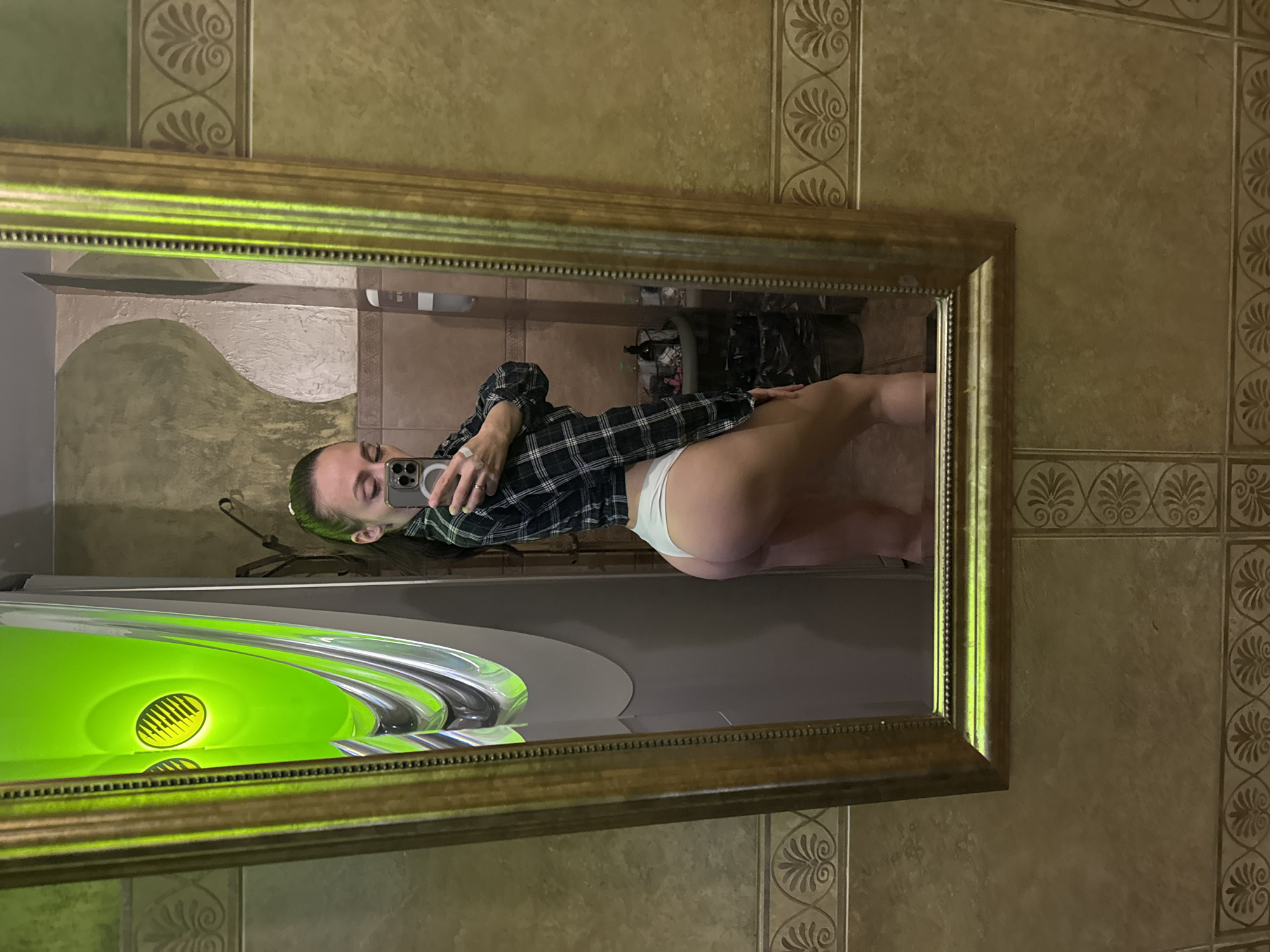 The image is a mirror selfie showing the reflection of a woman in a large, rectangular mirror with an ornate golden frame. The woman, likely standing upright while taking the photo, appears to be lying down in the rotated reflection. She has dark hair, possibly with a green tint, pulled back, and is holding a smartphone (likely an iPhone) to capture her image. She is wearing a dark plaid shirt and light-colored underwear. Her face is visible, looking towards the phone.
The background visible in the mirror's reflection features tiled walls with a decorative, scroll-like pattern, suggesting an indoor setting such as a bathroom or dressing room. To the left within the reflection, a striking, large, vibrant green object is visible, possibly a decorative vase or a light fixture, featuring an illuminated yellow oval pattern. A black trash bag or container is also discernible in the reflection behind the woman, along with a wall hanger or hook. The lighting is artificial and consistent with an indoor environment. No text is visible in the image. The overall impression is of a self-portrait taken in a personal, well-decorated interior space.