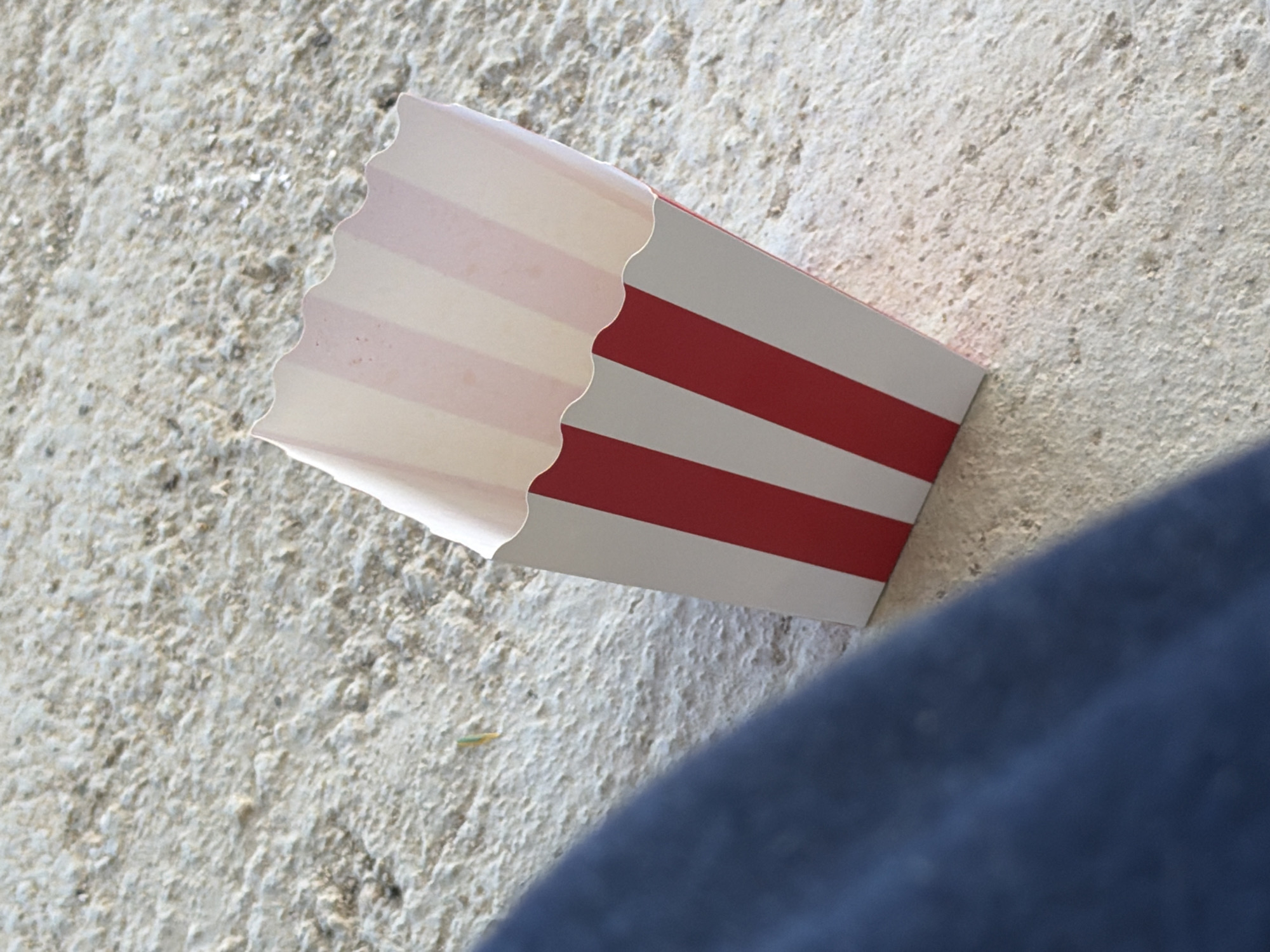 The image shows a close-up, high-angle shot of a red and white striped popcorn box resting on a textured, light-colored surface, likely concrete or pavement. A person's dark blue clothing is visible in the lower left corner of the frame, suggesting they are seated or leaning near the box. A small, thin object, possibly a piece of straw or grass, is visible on the ground to the left of the popcorn box. The background is a rough, granular texture of a light beige or gray color. The lighting suggests an outdoor environment, possibly in a public area like a park or street in Sant Vicenç de Montalt, Spain.