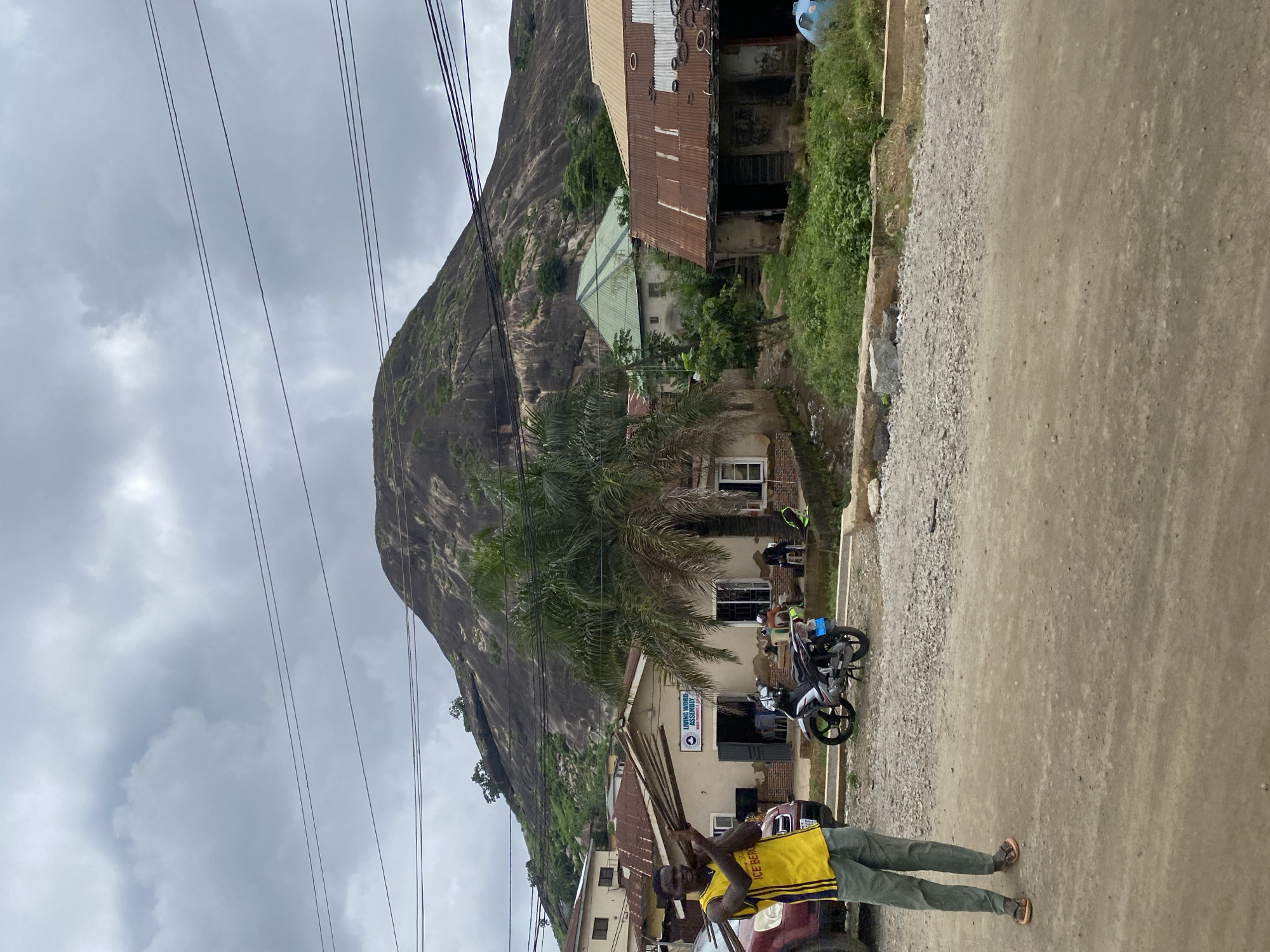 In Suleja, Nigeria, a man in a yellow vest and grey pants carries a bundle of wooden poles, walking on a dirt road in front of a building with a sign that reads "Living Word Assembly" and a palm tree. A motorcycle is parked nearby. In the background, a large, rocky hill with green vegetation dominates the skyline, with several buildings and power lines scattered below. The sky is overcast.