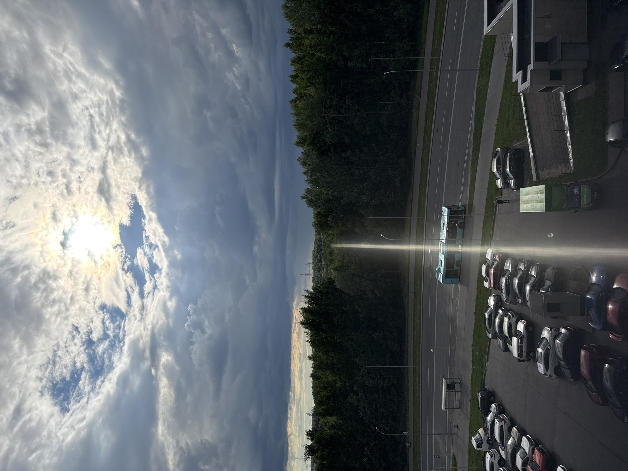 The image depicts a wide shot of a road with a light blue bus driving on it, surrounded by parked cars in a lot. In the background, there is a dense green forest, and the sky is filled with dramatic clouds, with the sun shining through a gap, creating a bright beam of light. The scene appears to be an urban or suburban setting, possibly on the outskirts of Saint Petersburg, Russia, given the presence of the forest bordering the road and parking lot. The lighting suggests it is either early morning or late afternoon, with the sun casting long shadows. There is no explicit indication of the exact location within Saint Petersburg.