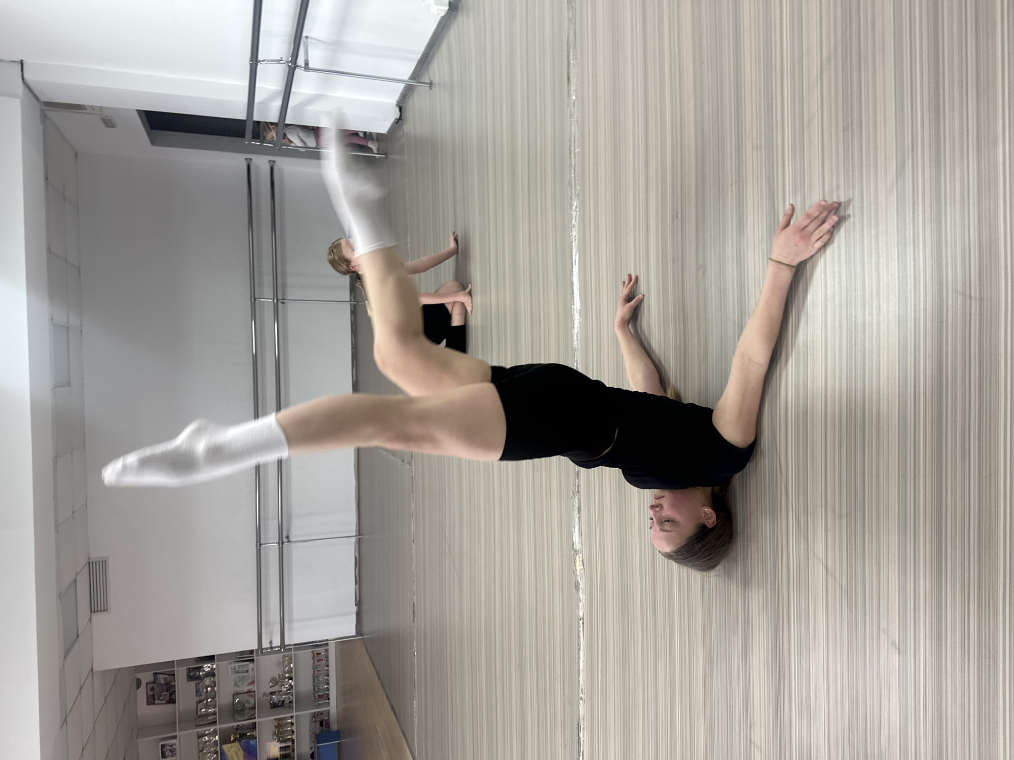 The scene is an interior view of a dance studio located in Votkinsk, Russia. In the foreground, a light-skinned female with medium-brown hair lies supine on a striped floor. She wears a black t-shirt, black shorts, and white socks, with her legs extended vertically and slightly bent at the knees. Her arms are extended laterally on the floor, and a thin dark bracelet is visible on her left wrist. To her left, in the mid-ground, a second light-skinned female with light hair kneels on the floor, also in dark attire and white socks, facing right. A third individual is partially visible behind a silver barre on the far right. The studio has white walls, a white drop-tile ceiling with an air vent, and multiple silver dance barres. A white shelving unit on the left wall displays numerous trophies and framed items. The floor is composed of alternating light gray and beige horizontal stripes. A dark open doorway is visible on the right.