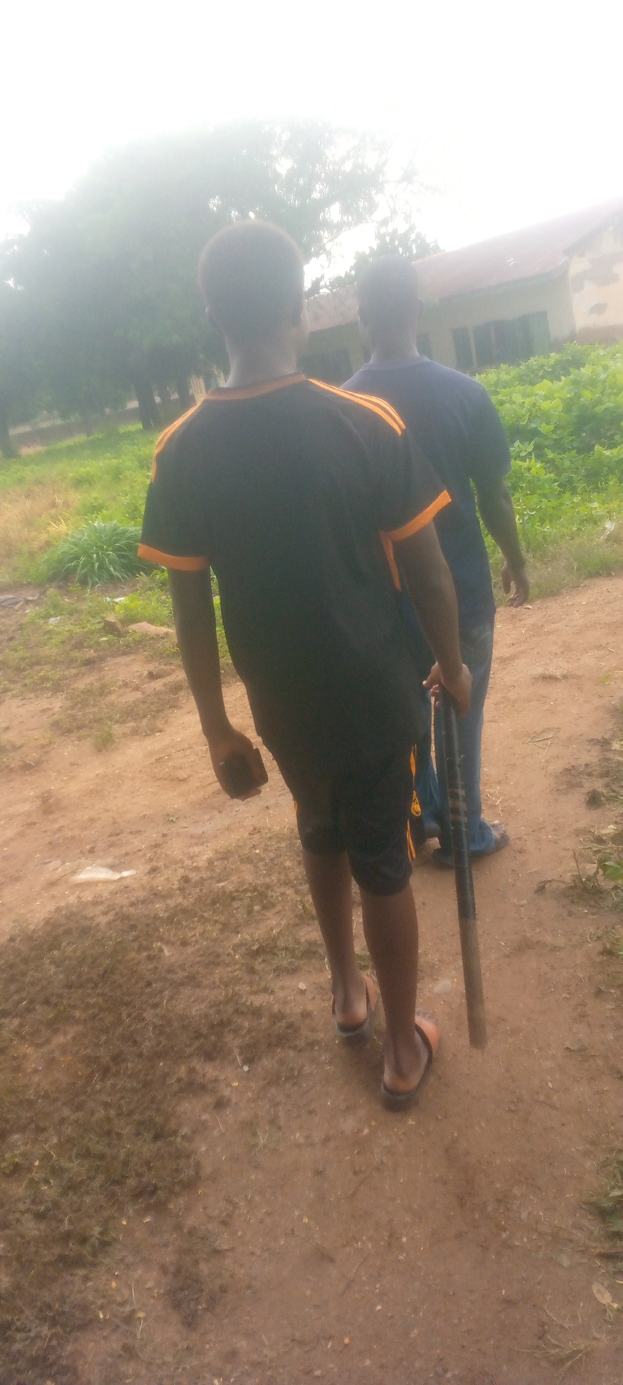Two individuals are shown from behind, walking on a dirt path. The person in the foreground is wearing a black t-shirt with orange stripes on the sleeves and collar, and black shorts. They are carrying a dark-colored object that appears to be a baton or stick in their right hand. The person behind them is wearing a dark blue t-shirt and jeans. They are walking slightly behind and to the right of the first person. The environment appears to be outdoors, possibly in a rural or semi-urban setting, with trees, vegetation, and a building visible in the background. The ground is unpaved and appears to be a dirt path with some patches of grass. The lighting suggests it is daytime, with overcast or hazy conditions.