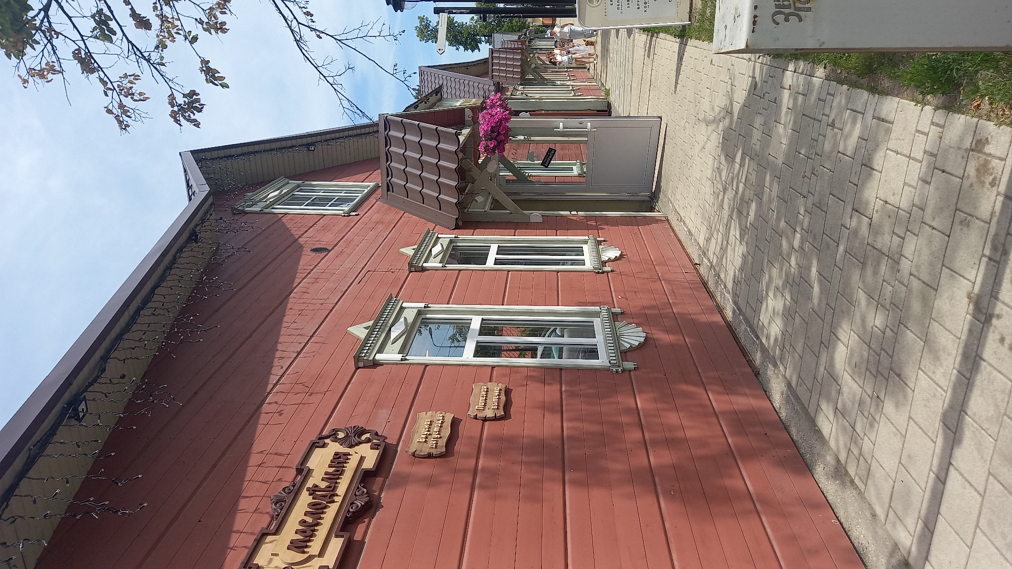 This image presents a ground-level street view in Zvenigorod, Russia, showcasing a row of traditional-style wooden buildings under a clear, bright sky. The primary building in the foreground and extending into the midground features reddish-brown horizontal plank siding. Its roof is dark brown, exhibiting a distinct tile pattern. Decorative strings of small, clear, icicle-like lights are suspended from the building's eaves.
The facade of the building is punctuated by multiple rectangular windows with white frames, each bordered by ornate light-green wooden trim. A large, carved wooden sign prominently displayed on the upper left portion of the building reads "МАСЛОДЕЛЬНЯ" (Maslodel'nya). Below this, two smaller, rectangular wooden signs are mounted vertically; the upper one states "Вкусная дегустация" (Delicious tasting), and the lower one "Полезная выпечка" (Healthy baked goods).
Several recessed doorways, each sheltered by a dark brown awning matching the roof material, are visible along the building. The closest doorway, positioned in the midground, features a light-grey door that is partially open. A hanging basket filled with vibrant pink flowers is suspended near this entrance.
A light-grey, square-paver sidewalk runs parallel to the building, exhibiting distinct shadows from unseen overhead foliage. Further along the sidewalk, multiple individuals, primarily women in casual attire, are observed walking away from the viewer. A dark utility pole or signpost with attached directional arrows is situated on the right side of the sidewalk in the midground. A white display stand or sign is partially visible in the bottom right corner of the frame. In the upper right, a bare tree branch with sparse leaves extends into the sky.