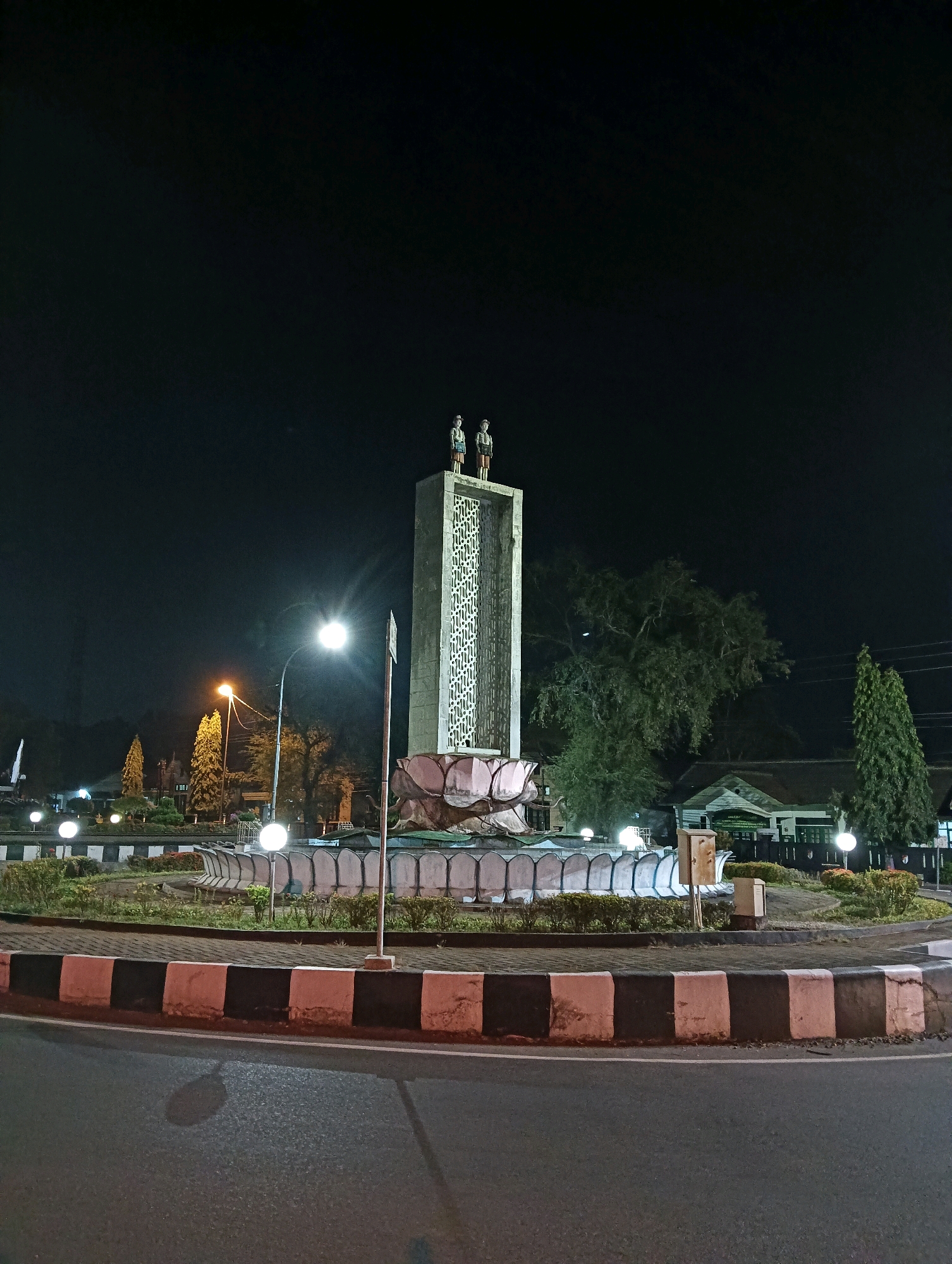 The image shows a monument at night in a roundabout in Purworejo, Indonesia. The monument is a tall, rectangular structure with a patterned facade, topped by two standing figures. At the base of the monument is a circular fountain with lotus flower-shaped elements. The roundabout is illuminated by streetlights, and there are decorative lights around the fountain. Trees and buildings are visible in the background. The foreground shows the asphalt road and the curb of the roundabout, which is painted with alternating black and white stripes.