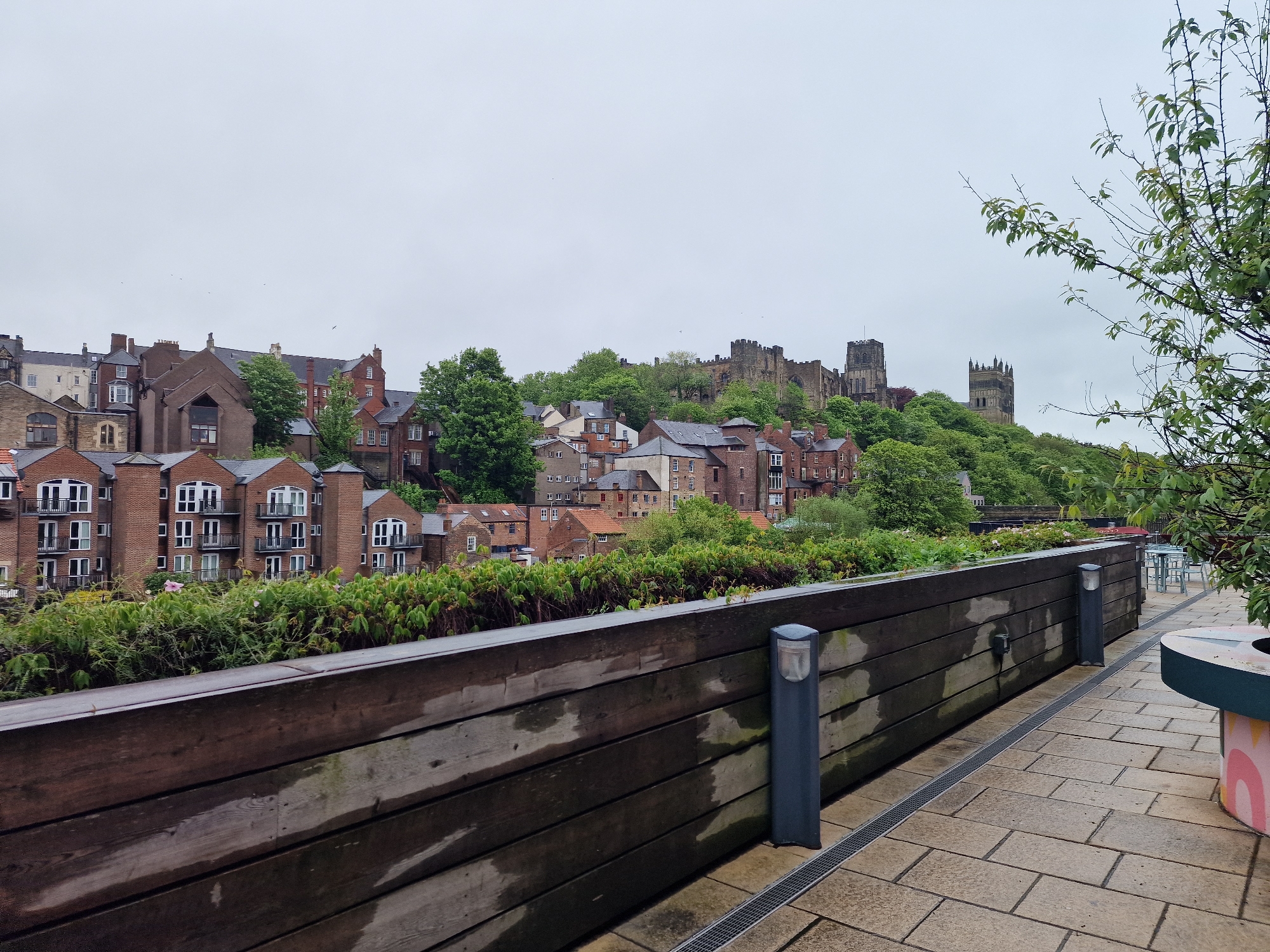 The image depicts a view of Durham, United Kingdom, from an elevated walkway or platform. In the foreground, there is a wooden barrier with metal posts and integrated lights, with a paved walkway to the right. A tree or large shrub is on the right side of the walkway. Beyond the barrier, there are several buildings of various architectural styles, mostly constructed of brick with slate or tiled roofs. A few trees are interspersed among the buildings. In the background, Durham Cathedral and Durham Castle, are visible on a hill. The sky is overcast and gray. No people are visible in the image.