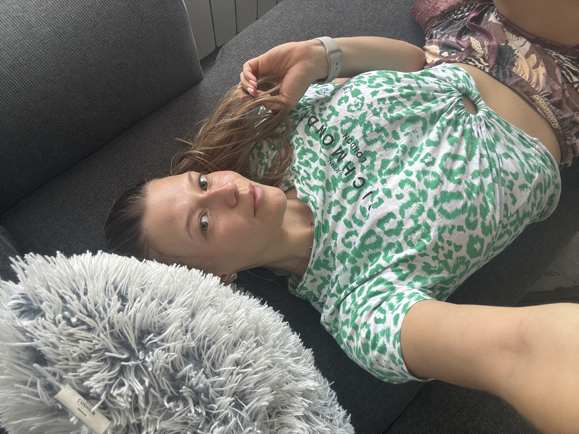 A woman lies on a grey sofa, her body angled towards the camera. She is wearing a green and white leopard-print t-shirt tied at the front, exposing her midriff, and patterned shorts. To her left is a fluffy white cushion. The scene appears to be an indoor setting, likely a living room. There are no explicit indications of the location being in Sofia, Bulgaria.