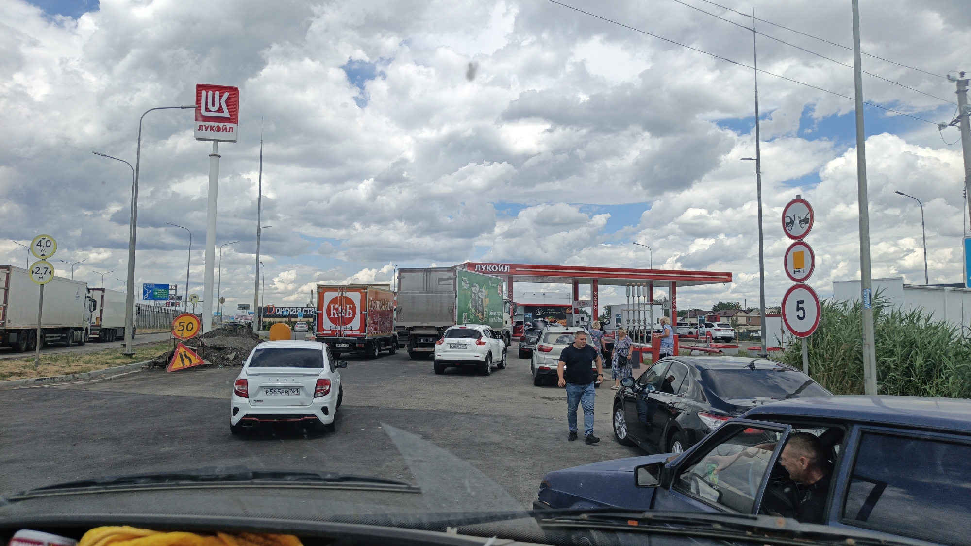 The image depicts a traffic scene at a Lukoil gas station on a cloudy day. Several vehicles, including trucks, cars, and a white Lada sedan, are present. Some vehicles are queuing at the gas pumps. People are visible near the gas station and some are standing outside their cars. There are traffic signs indicating height restrictions (4.0 m and 2.7 m), a speed limit of 50 km/h, and other regulatory signs. The environment appears to be an open area with some vegetation and buildings in the background. The overall impression is that of a busy commercial area with a moderate flow of traffic.