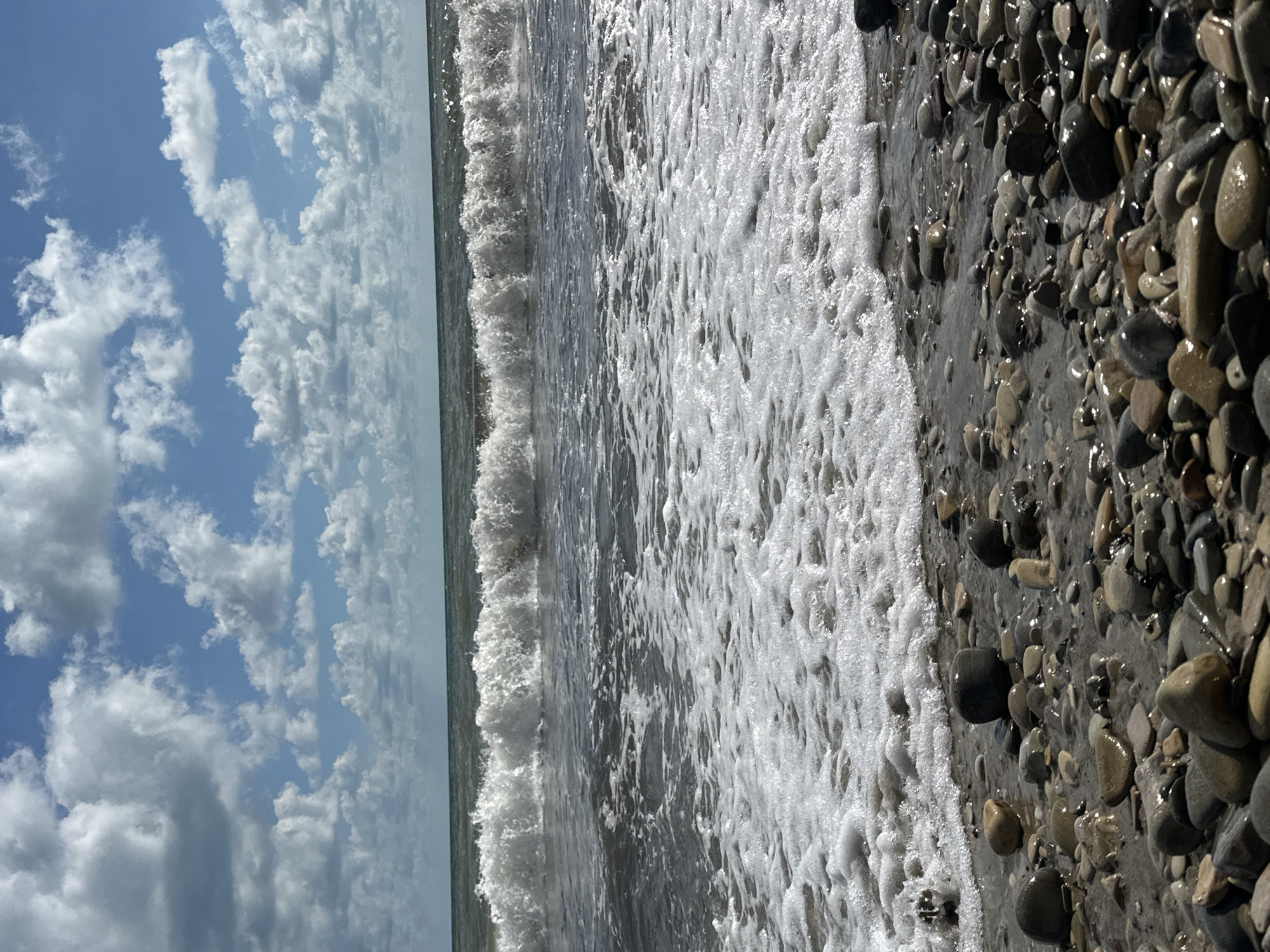 The image depicts a rocky beach on a partly cloudy day. The foreground is filled with smooth, wet pebbles of various sizes and colors, interspersed with patches of wet sand. A line of white foam from a receding wave marks the edge of the water. In the middle ground, a wave is breaking with white crests and turbulent water. The background shows the dark, choppy sea stretching to the horizon, under a sky with scattered white cumulus clouds against a blue backdrop. The location is identified as Lermontovo, Russia. There are no people visible in the scene.