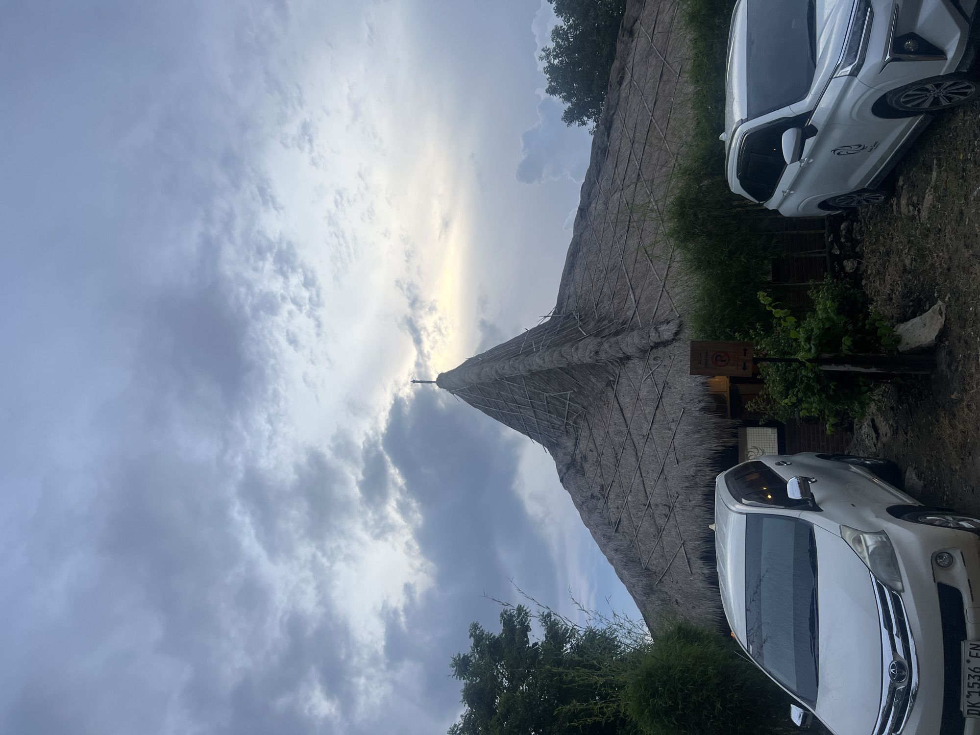 The image depicts an outdoor scene in what appears to be Nusa Penida, Indonesia, featuring a large building with a distinctive thatched roof and two white parked cars under an overcast sky.
The prominent subject is a tall, conical building, likely constructed from natural materials, with a intricately thatched roof that slopes steeply upwards to a pointed apex. The texture of the thatch is visible, showing a grid-like pattern. Sunlight, possibly from a late afternoon or early morning sun, peeks through the heavy cloud cover, illuminating the peak of the roof and casting a soft glow on parts of the sky.
To the right of the building, two white multi-purpose vehicles (MPVs) are parked. The lower car is a Toyota, and its license plate "DK 1536 FN" is clearly visible. The upper car also has a dark, swirl-like logo on its passenger side door. Both cars appear to be clean and well-maintained.
The ground around the cars and building is unpaved and somewhat uneven, with areas of sparse greenery, dirt, and rocks. Bushes and trees with dark green foliage are visible behind the cars and in the lower left corner, contributing to the natural, rustic setting. A small, dark sign near the front of the lower car displays a red circular symbol with a diagonal line, typically indicating a prohibition, though the specific text or image within the circle is obscured. The sky is largely cloudy, suggesting either an overcast day or the transition period of dusk or dawn. No specific human activity is evident in the scene.